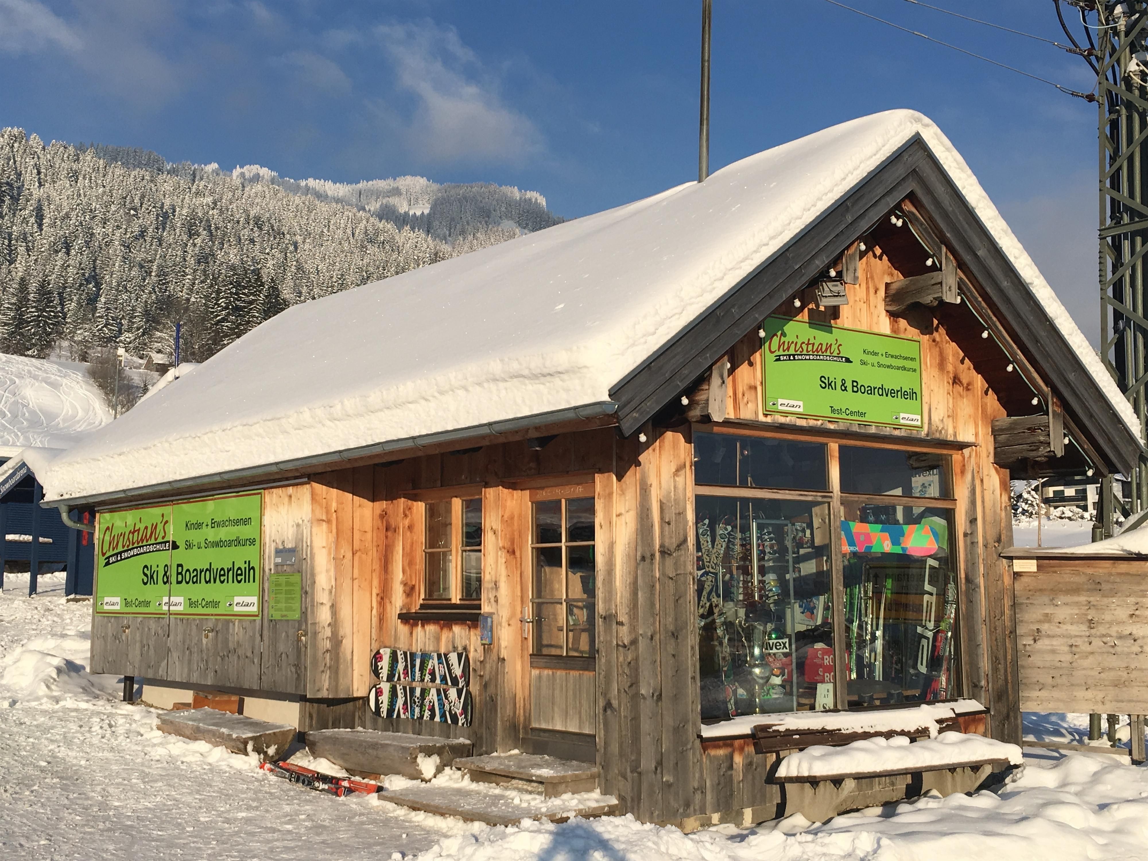 Ein gemütlicher Skiverleih aus Holz mit einem schneebedeckten Dach. Im Hintergrund sind verschneite Berge zu sehen.
