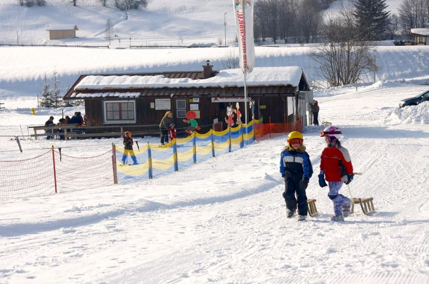 Zwei Kinder mit Helmen und Skikleidung ziehen einen Holzschlitten über eine verschneite Fläche.