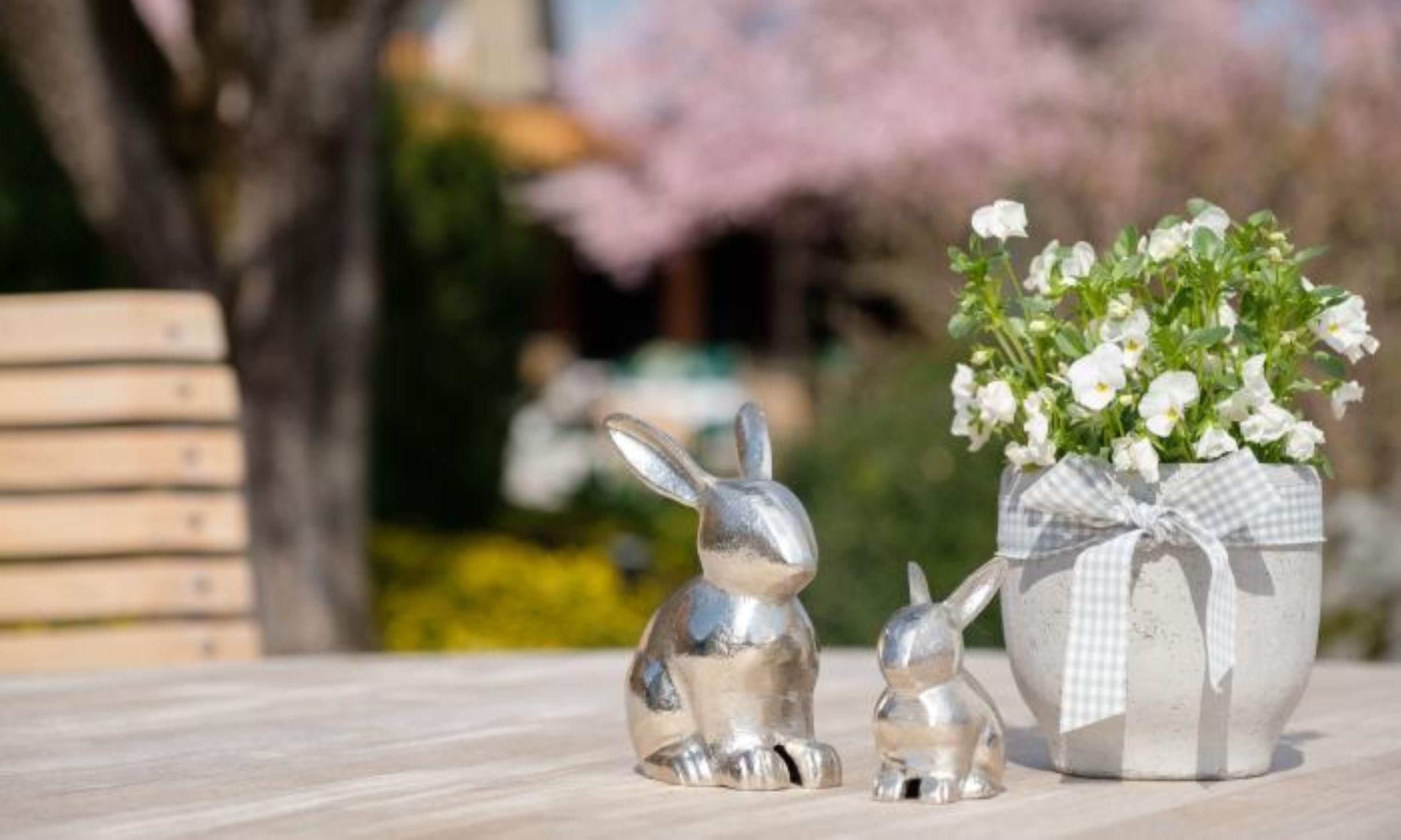 Zwei silberne Deko-Osterhasen und ein Blumentopf auf einem hölzernen Tisch.