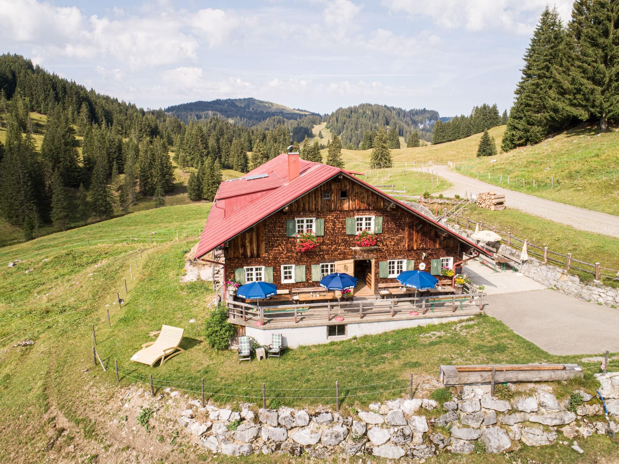 Eine charmante Alpe in der Landschaft, umgeben von grünen Wiesen und Bergen. Die Veranda ist mit sonnigen Stühlen und bunten Blumen dekoriert.