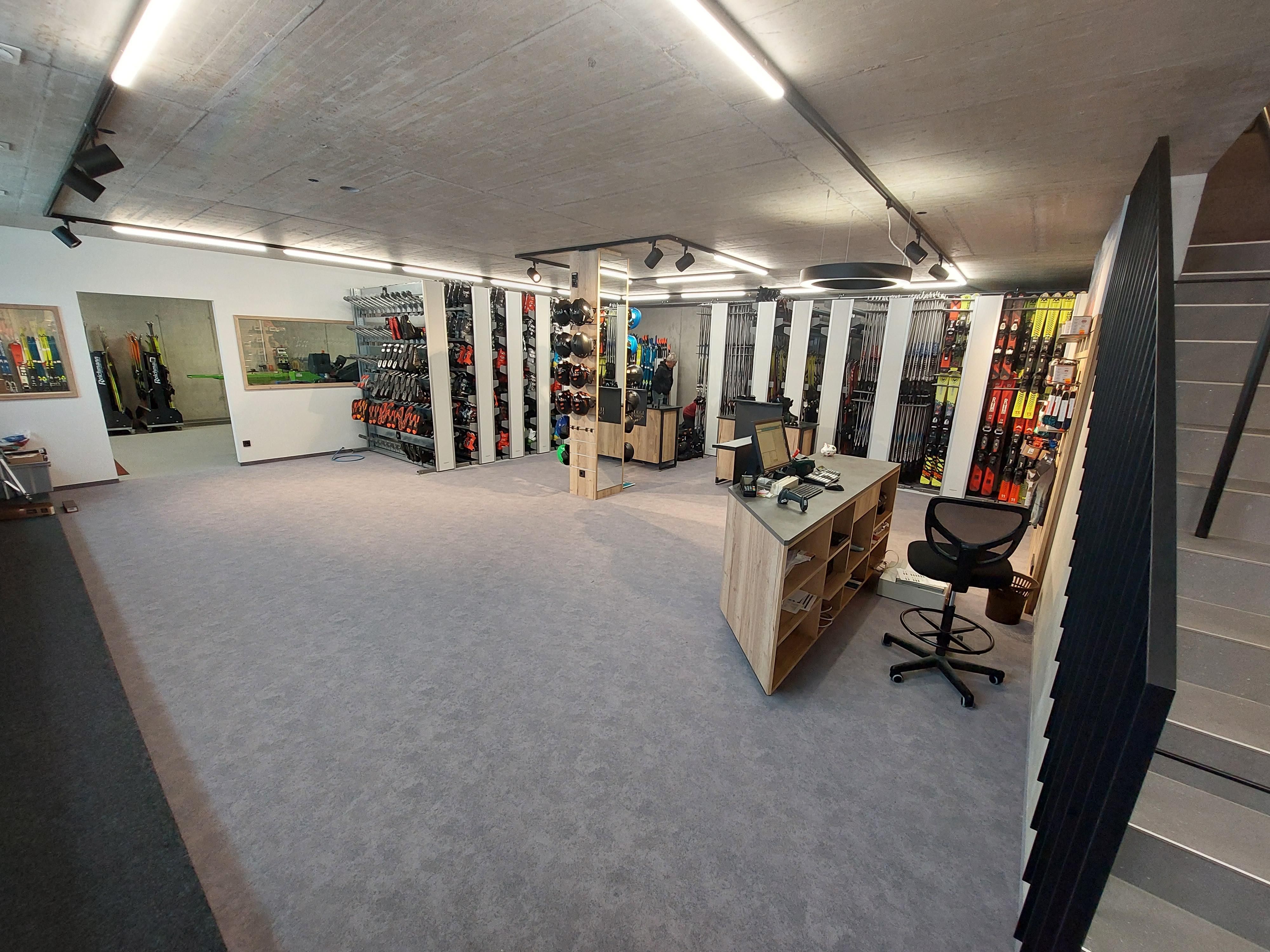 Sportgeschäft Luggi Endrös. Grauer Teppichboden, helle Decke mit Spots. Regale mit Skischuhen und Skiern. Kasse mit Computer und Bürostuhl. Treppenaufgang.