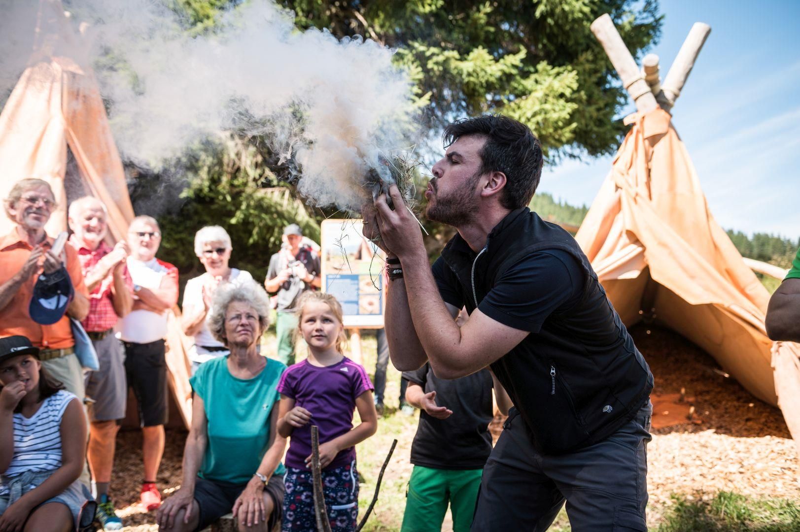 Ein Mann bläst Rauch in eine dunkle Masse vor einer Gruppe von Zuschauern. Zelte und Bäume im Hintergrund.