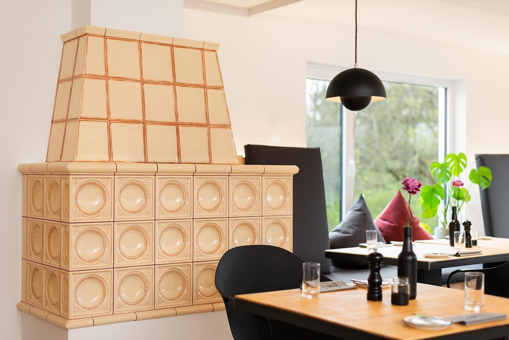 Kachelofen in Beige und Orange. Schwarzer Holztisch mit grauen Stühlen, Gläsern und Weinflaschen. Helle Wände, großes Fenster mit Blick ins Grüne. Schwarze Hängelampe.