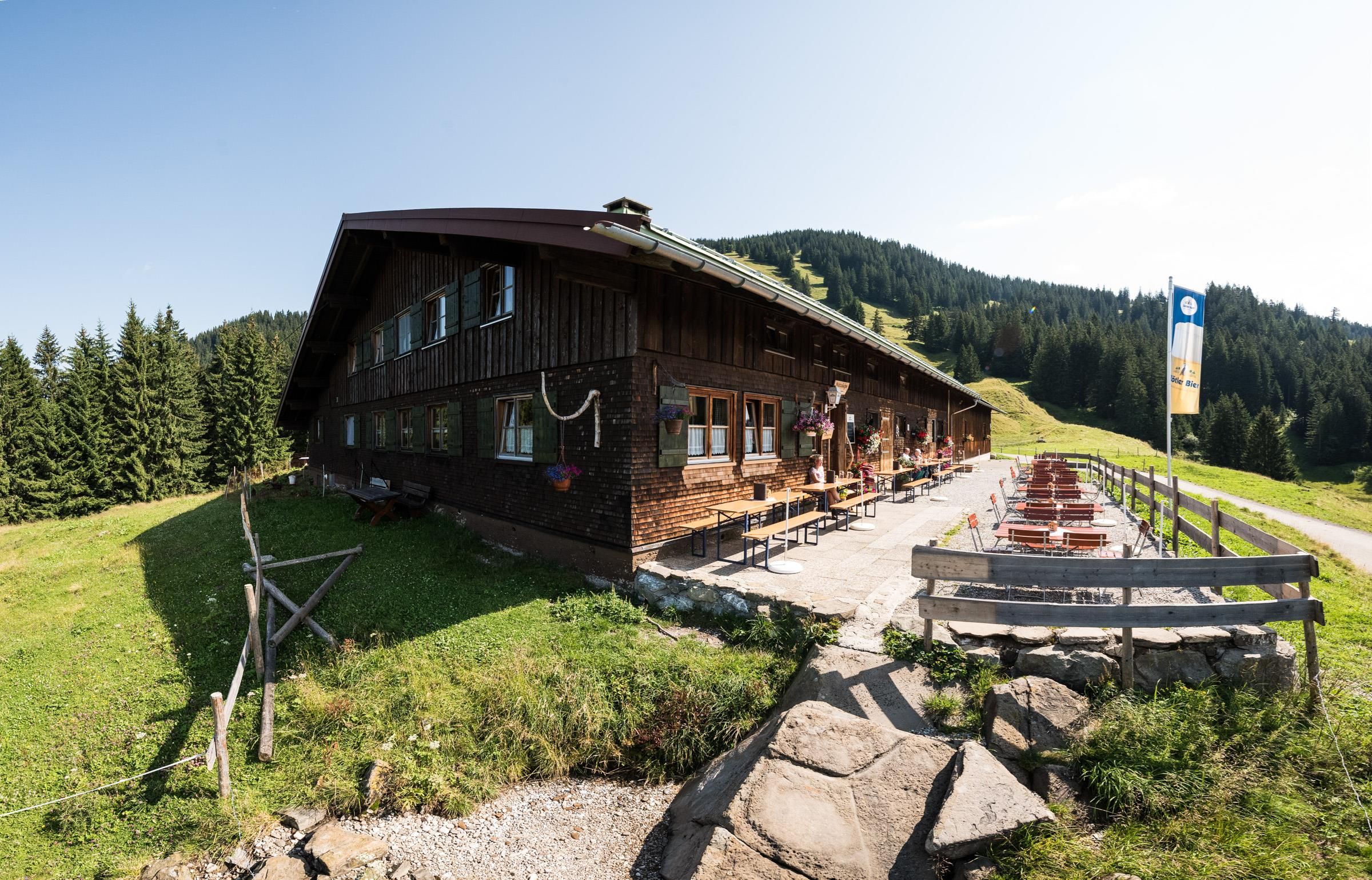 Dunkelbraunes Holzhaus mit grünen Fensterläden, lange Terrasse mit Tischen und Bänken, grüne Wiese und Bäume im Hintergrund, blauer Himmel.