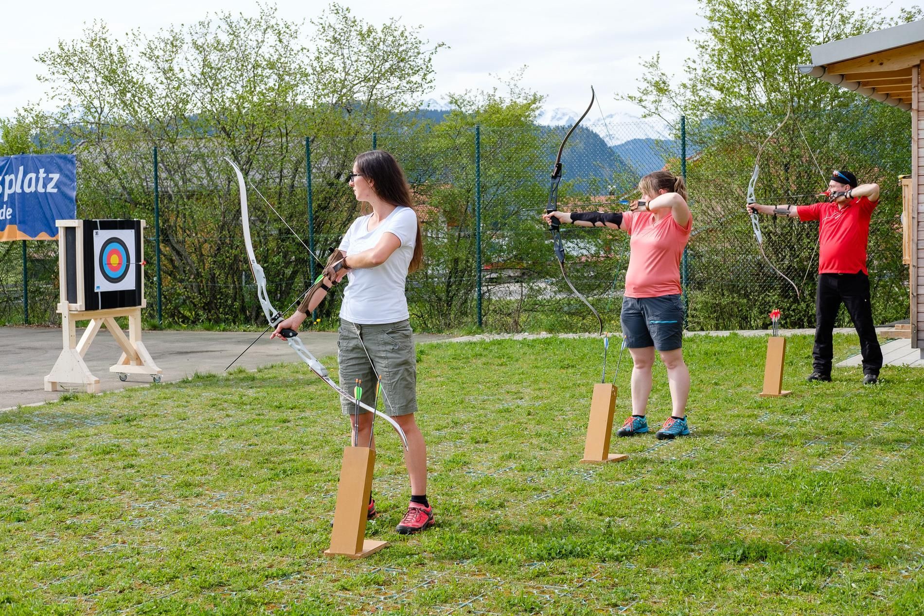 Drei Bogenschützen auf grünem Platz vor Zielscheibe.