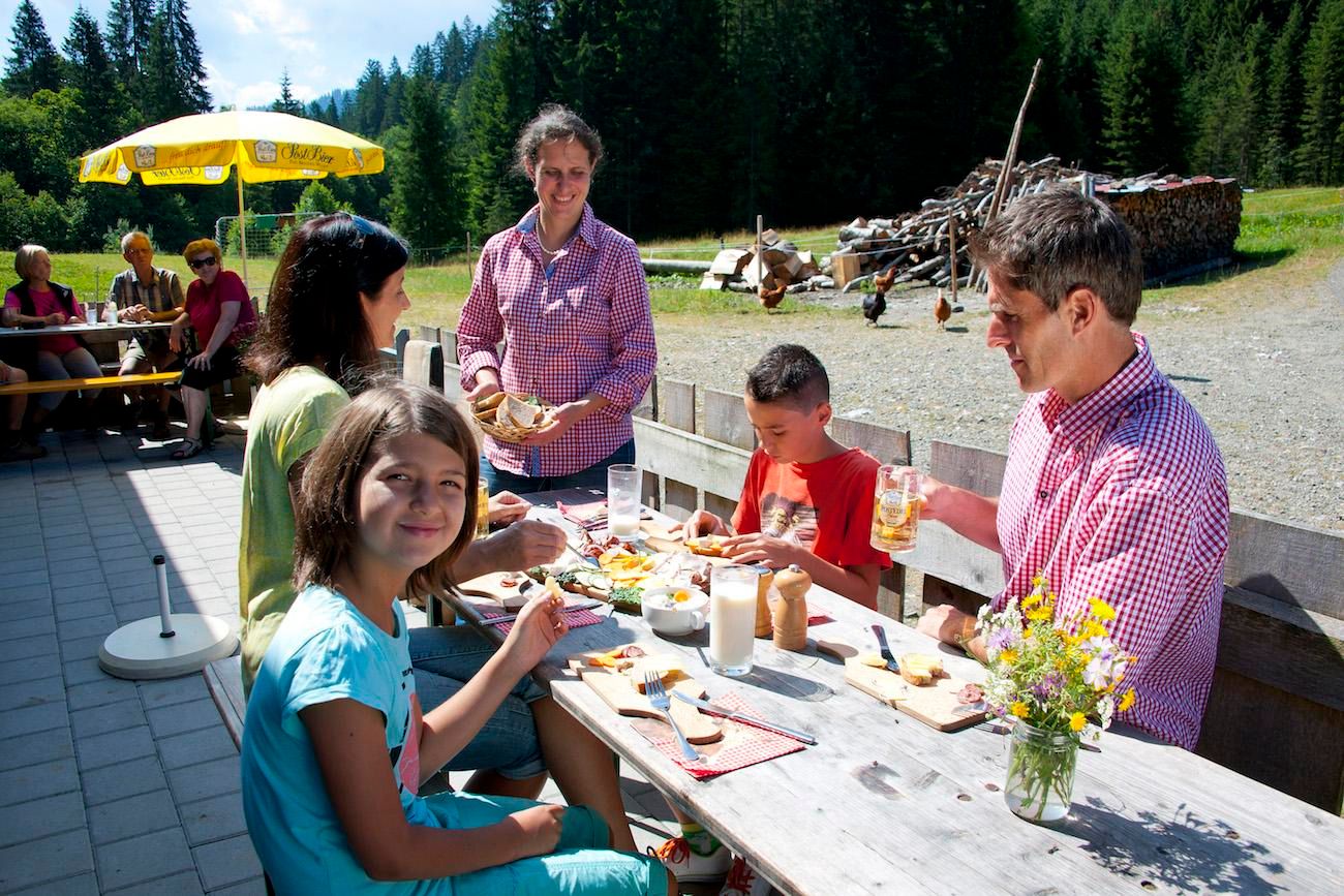 Familie isst auf der Lenzenalpe im Freien. Holztisch mit Brotzeit, Getränken und Blumen. Sonnenschirm spendet Schatten. Grüne Wiese und Bäume im Hintergrund.