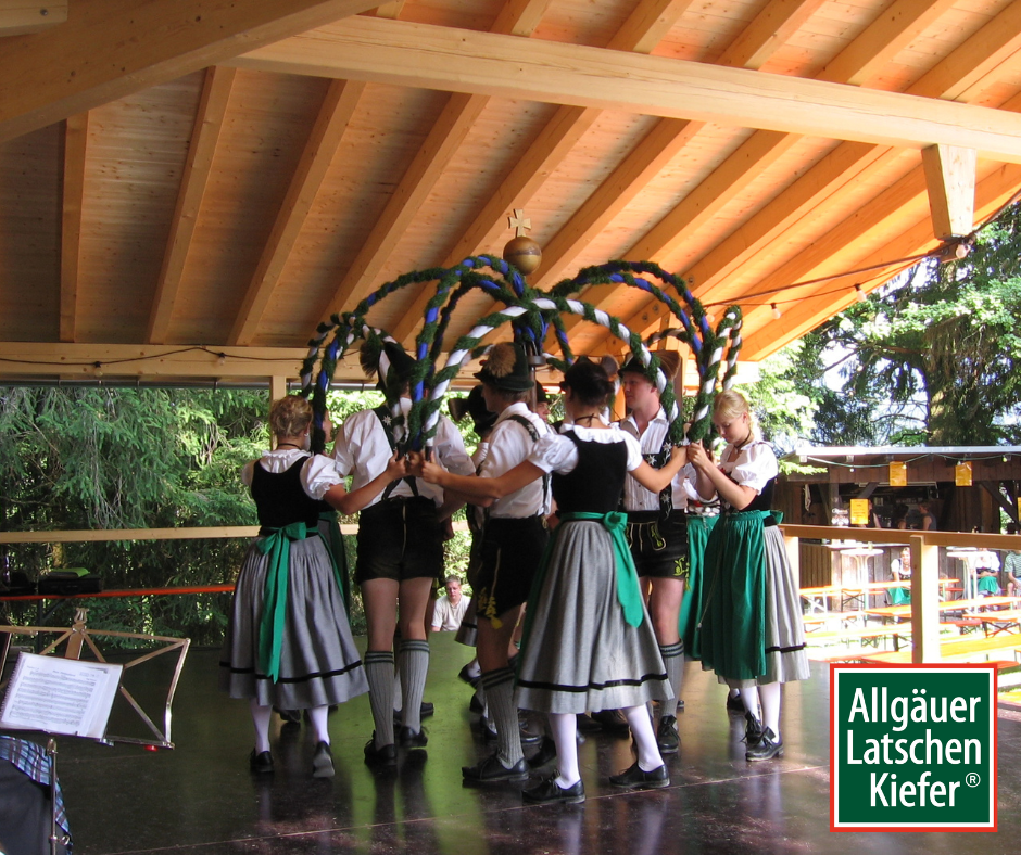 Acht junge Menschen in Tracht tanzen um einen Maibaum auf dem Waldfest in Obermaiselstein. Sie halten bunte Bänder. Holzbalkendecke über ihnen.