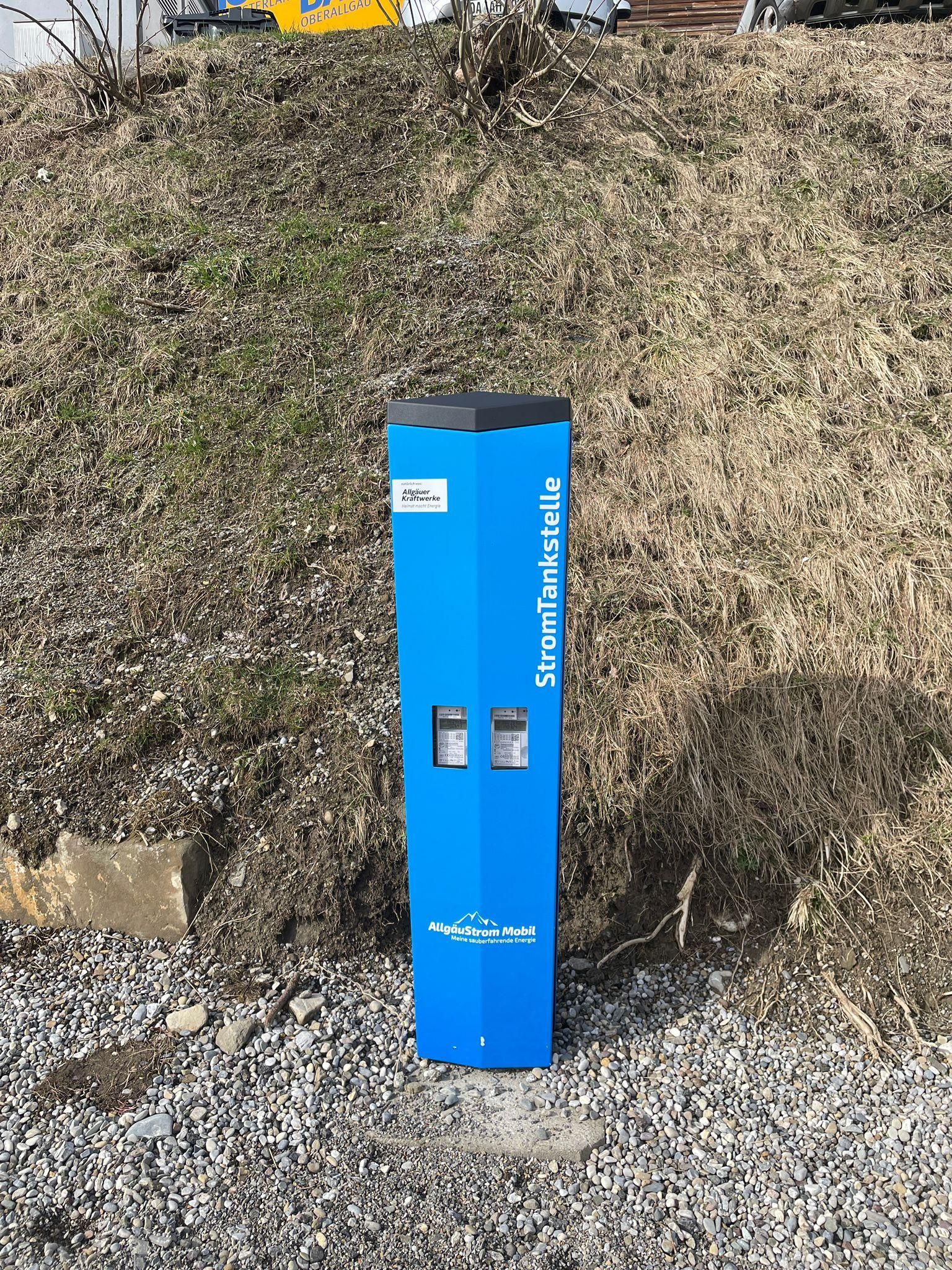 Eine blaue Stromtankstelle mit zwei Ladepunkten steht auf einem Schotterplatz vor einem grasbewachsenen Hang.