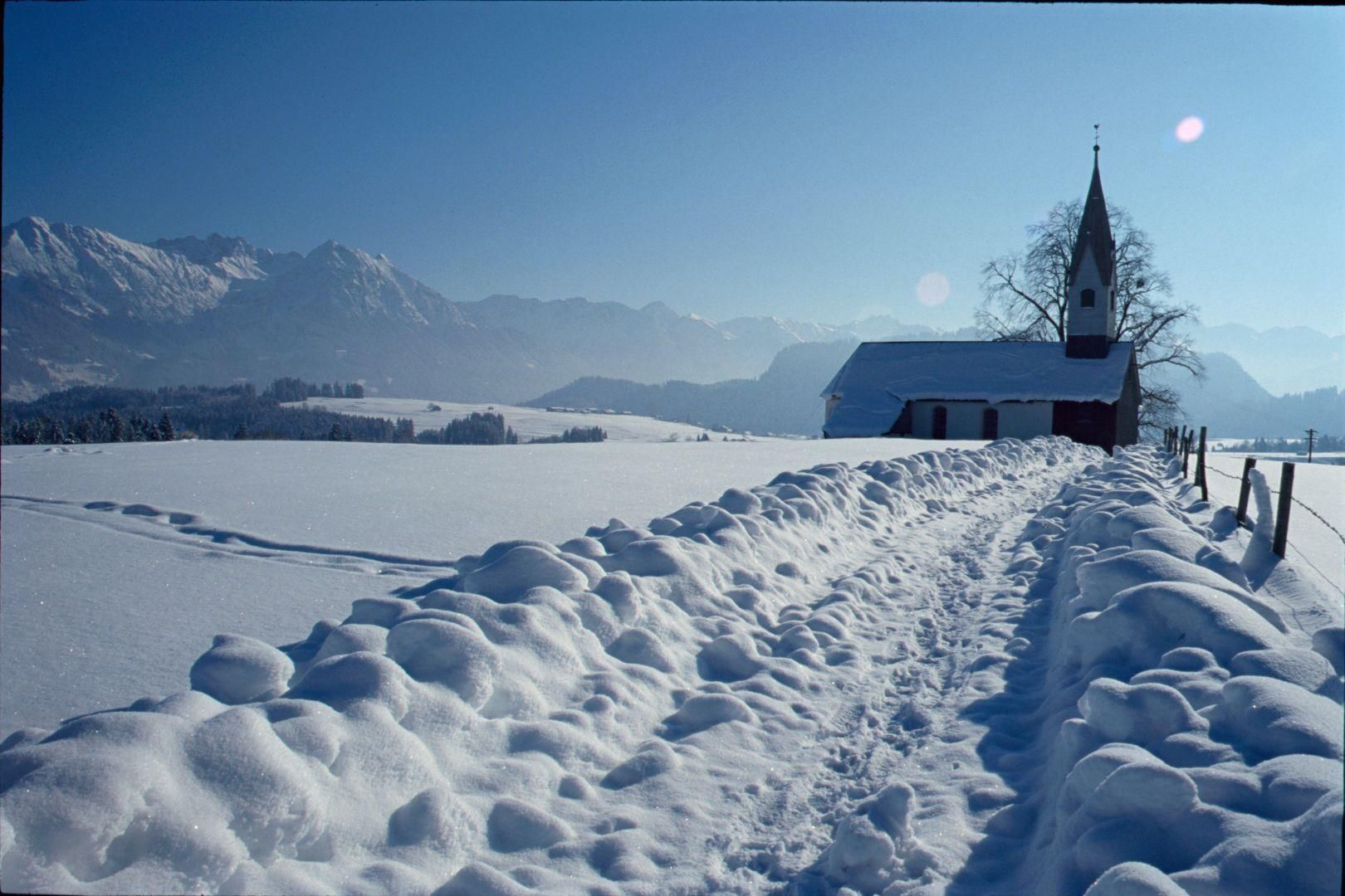 Weitläufiges, schneebedecktes Feld, rechts eine kleine Kirche mit spitzem Turm, dahinter Berge.