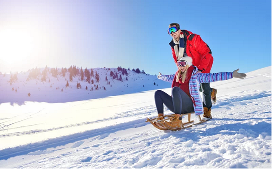 Ein fröhliches Paar rodet einen verschneiten Hügel hinunter, im Hintergrund sonnenbeschienene Winterlandschaft.