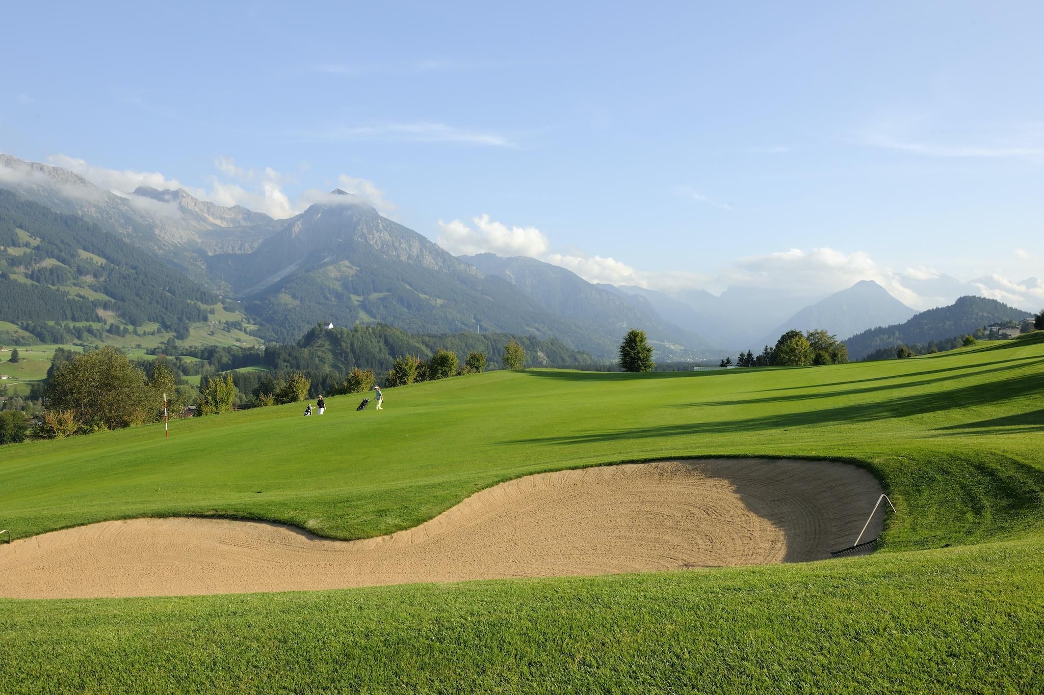 Hügeliger, grüner Golfplatz vor einer beeindruckenden Bergkulisse. Ein Sandbunker im Vordergrund.