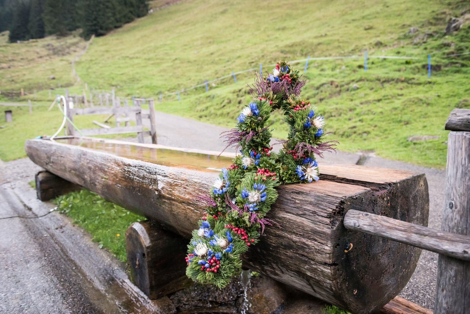 Ein Kranz aus Tannenzweigen und bunten Blumen ist an einem Baumstamm befestigt.