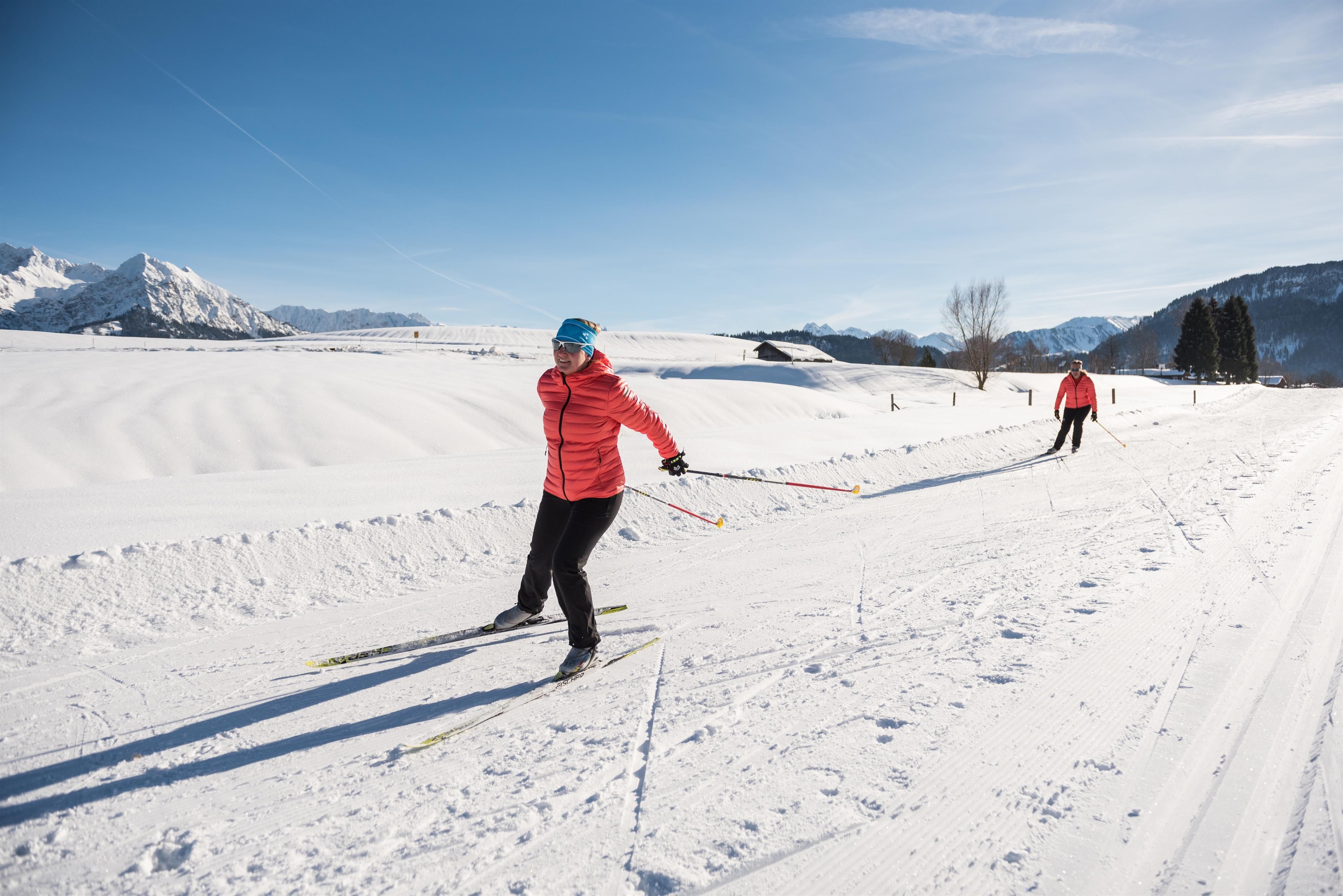 Sonnige Winterlandschaft: Zwei Langläuferinnen auf gespurter Loipe, Berge im Hintergrund.
