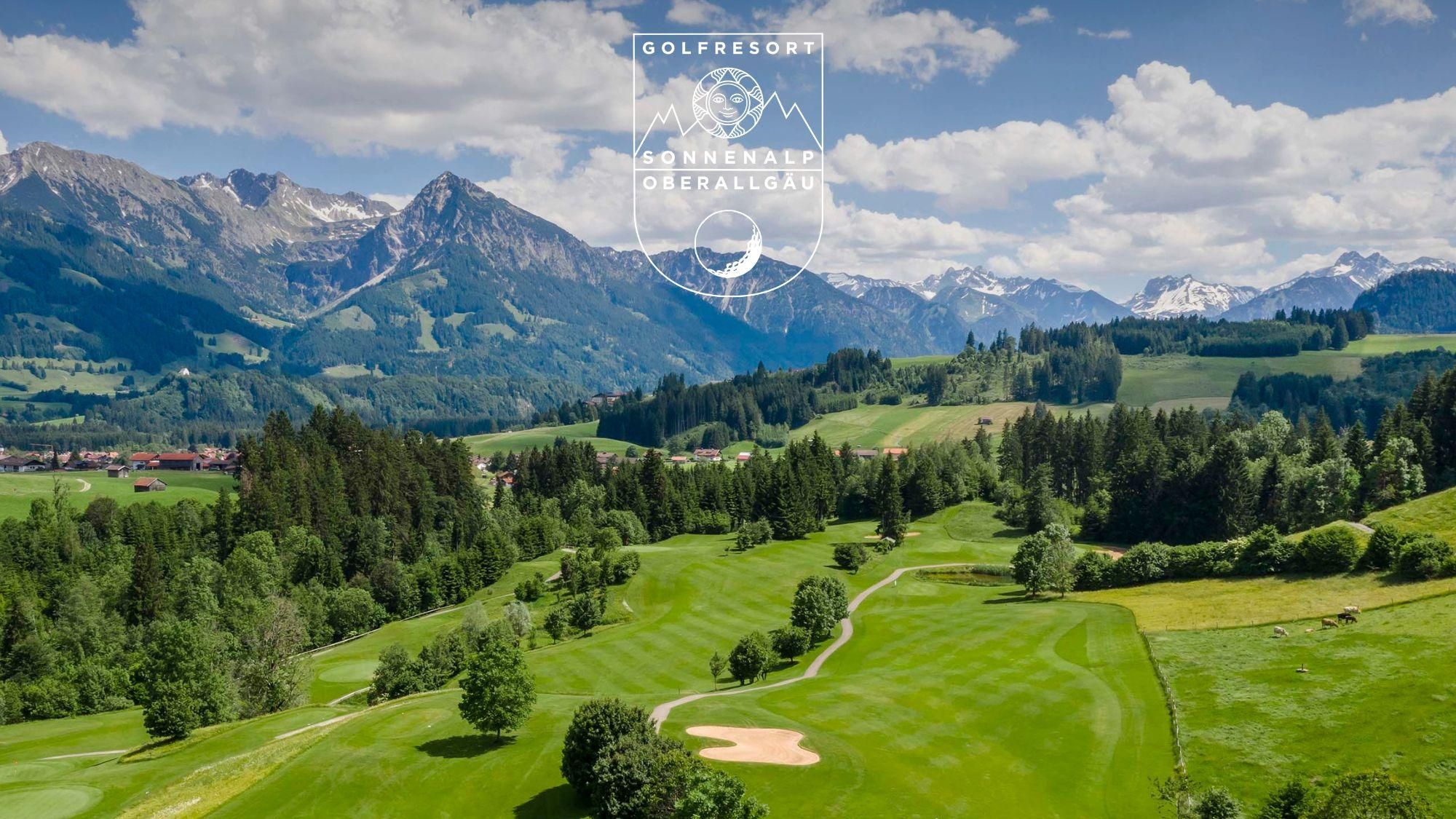 Weitläufiger, grüner Golfplatz Oberallgäu mit sanften Hügeln. Gepflegte Rasenflächen und einzelne Bäume. Im Hintergrund sind Berge unter einem hellen Himmel sichtbar.