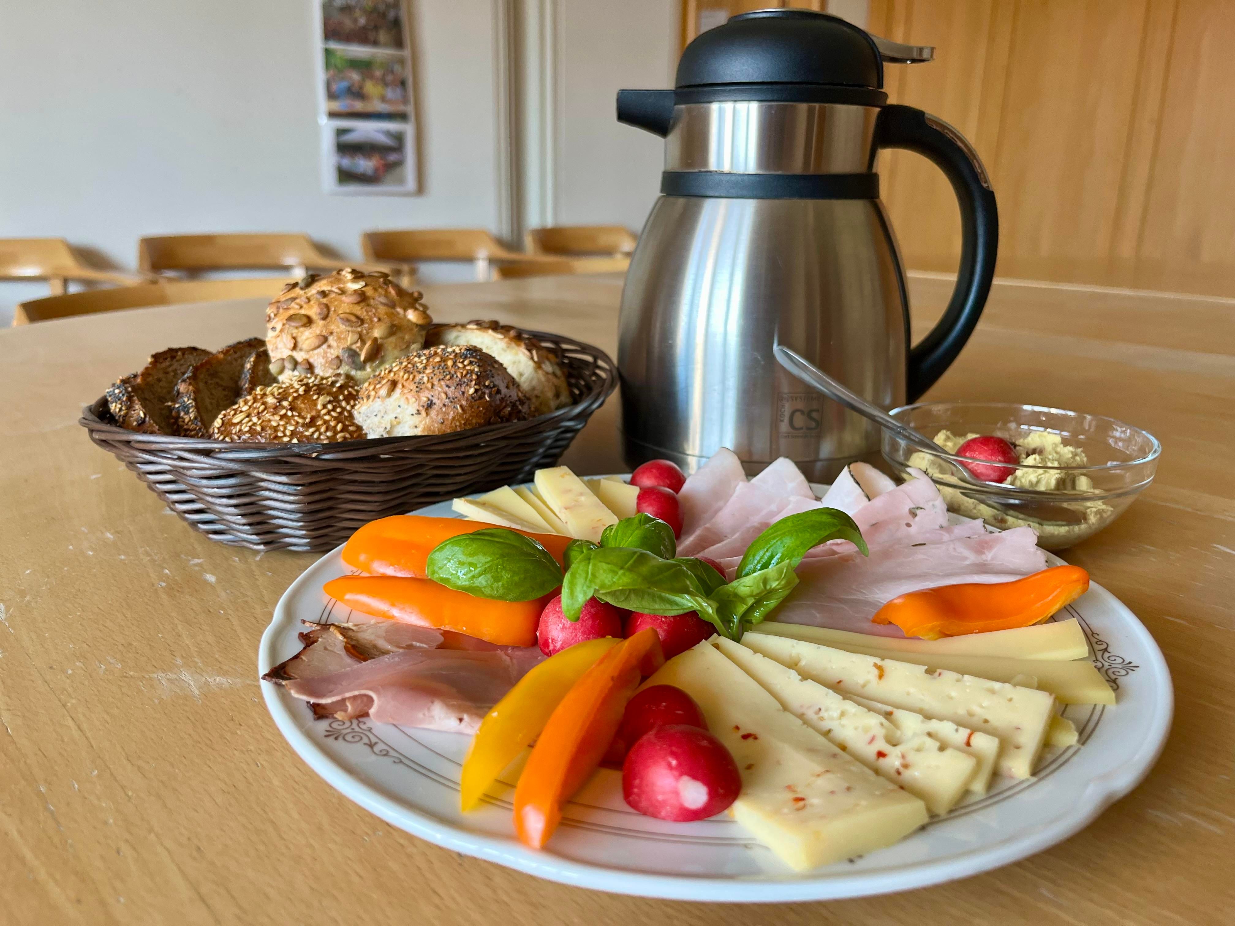 Ein Teller mit Käse, Aufschnitt und frischem Gemüse. Neben dem Teller steht eine Kaffeekanne und ein Korb mit Semmeln.