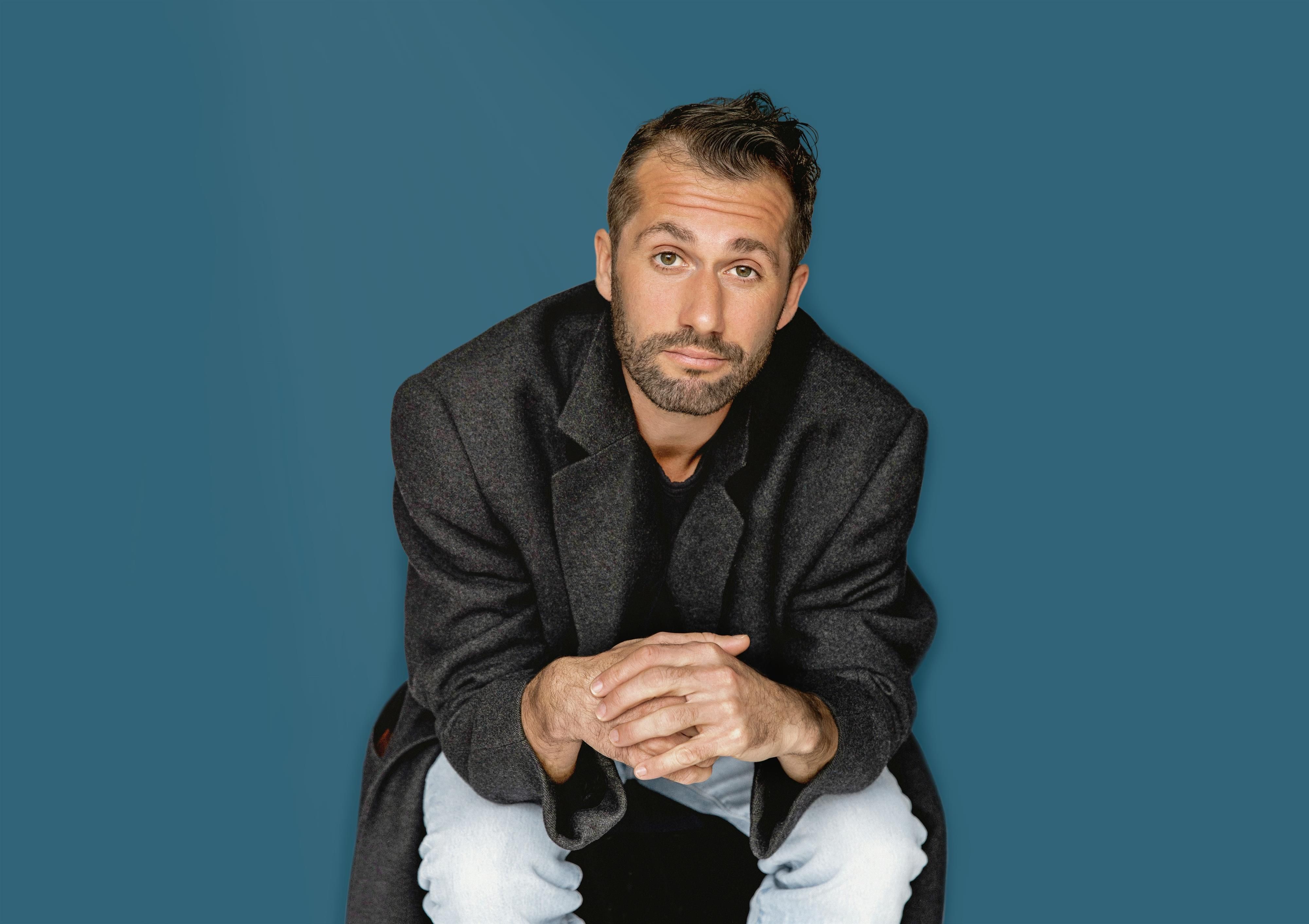 <p>Stefan Leonhardsberger in schwarzem Jacket und hellblauer Jeans - kniend in halbsitzender Position mit gefalteten Händen.</p>