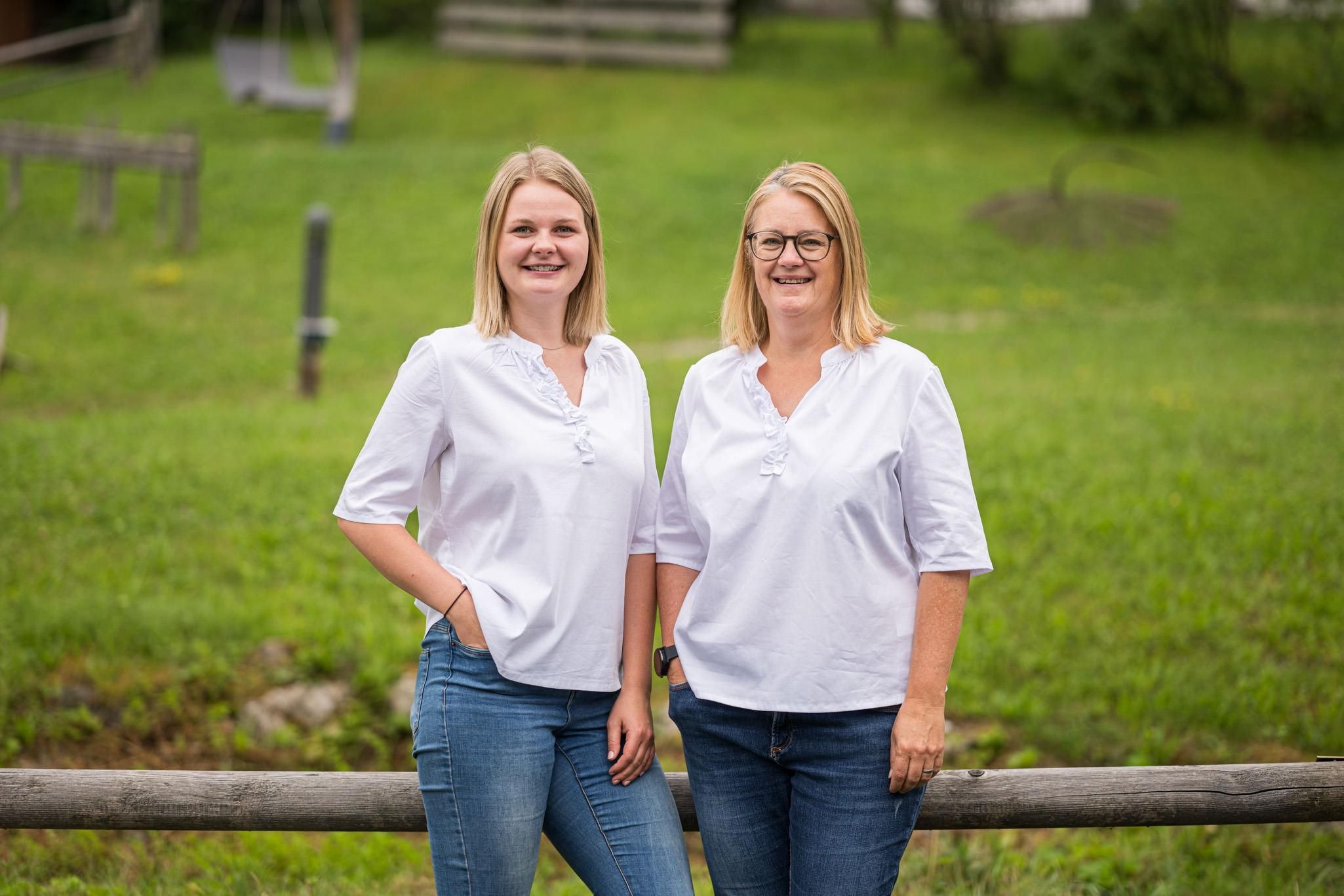 Zwei lächelnde Frauen in weißen Oberteilen und Jeans stehen vor einer grünen Wiese.