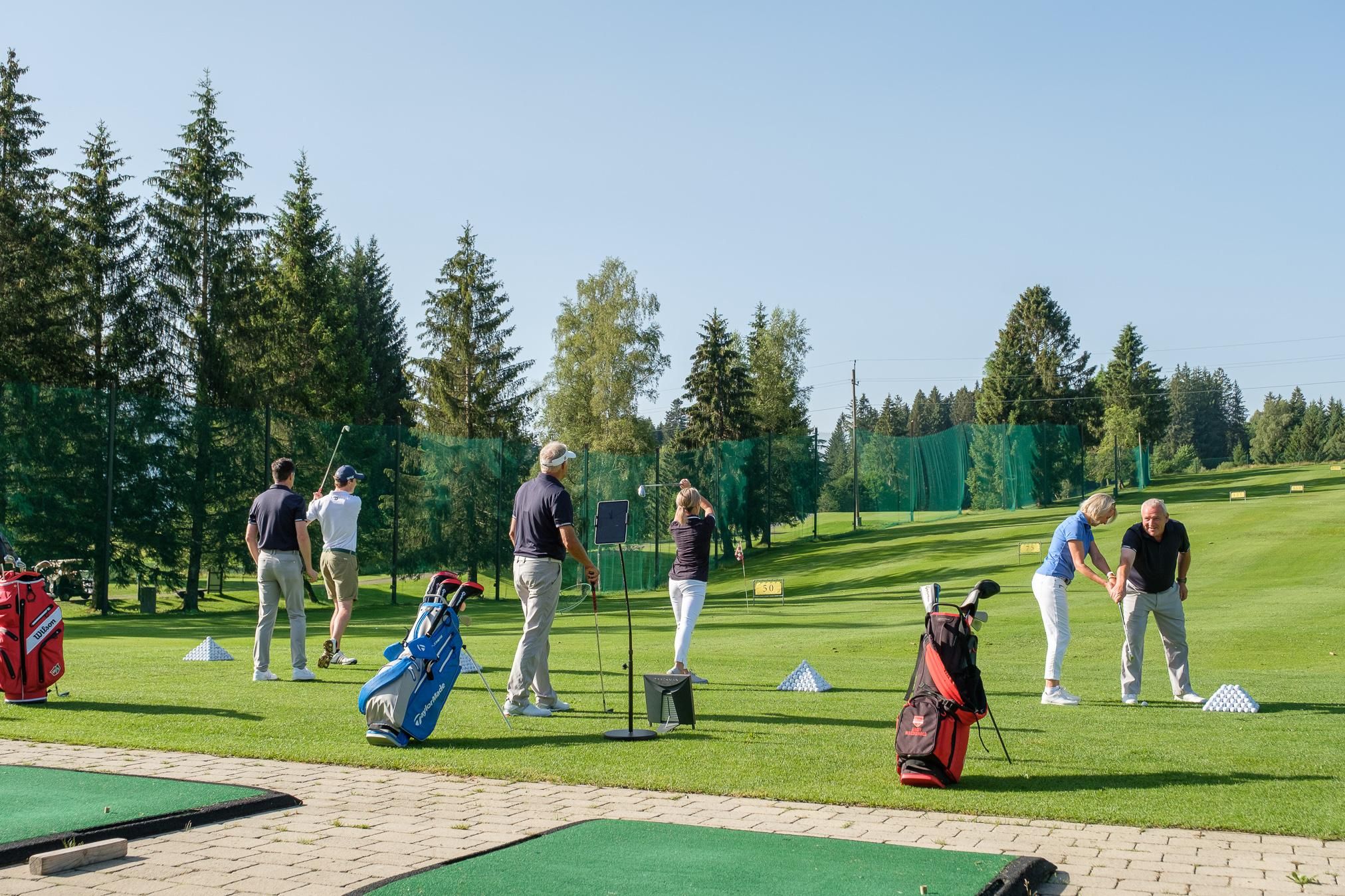 Mehrere Personen spielen Golf auf einem grünen Platz, umgeben von Bäumen unter blauem Himmel.
