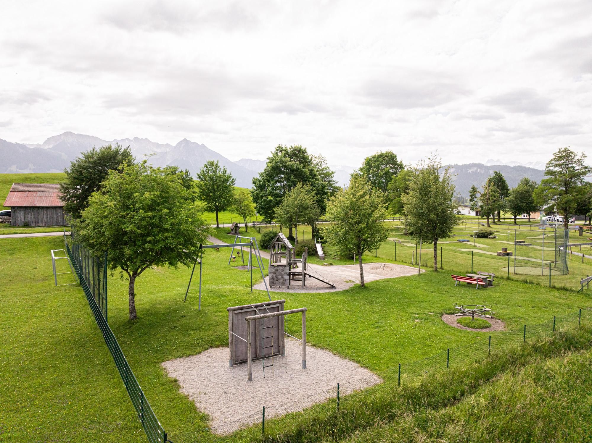 Ein schöner Spielplatz in einer grünen Umgebung mit Bäumen. Im Hintergrund sind Berge und ein weiter Himmel zu sehen.