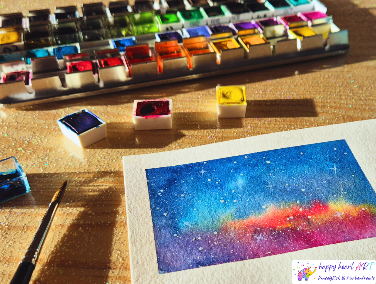 Viele Aquarellfarben von gelb bis schwarz auf einem Tisch, im Vordergrund ein Aquarellbild in Blau und Rottönen.