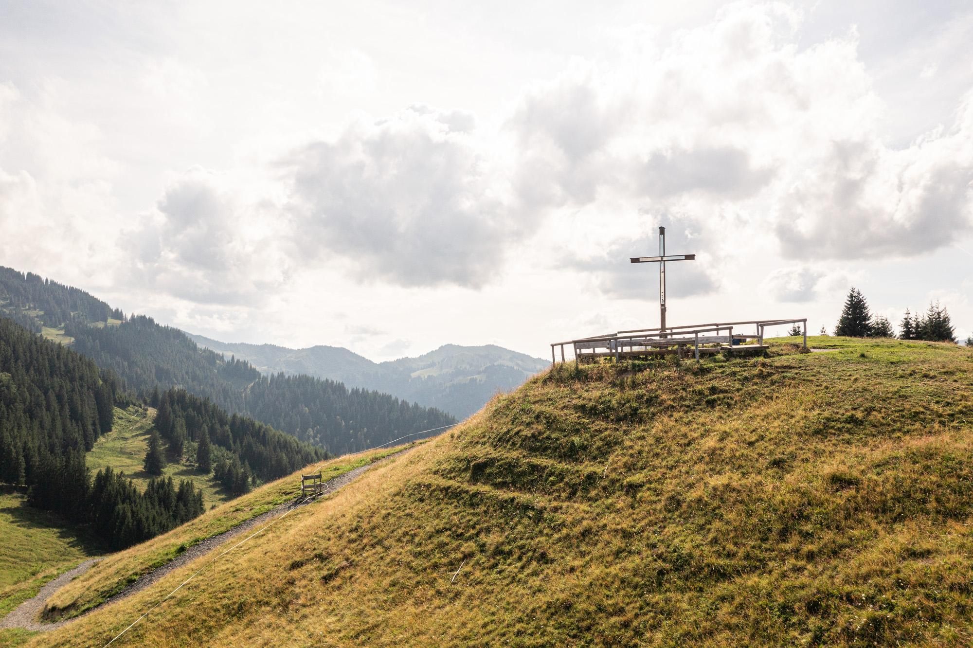 Eine sanfte Berglandschaft mit einem Holzkreuz am Ofterschwanger Horn und Blick auf die Berge. Der Himmel ist bewölkt und die Umgebung ist grün und ruhig.