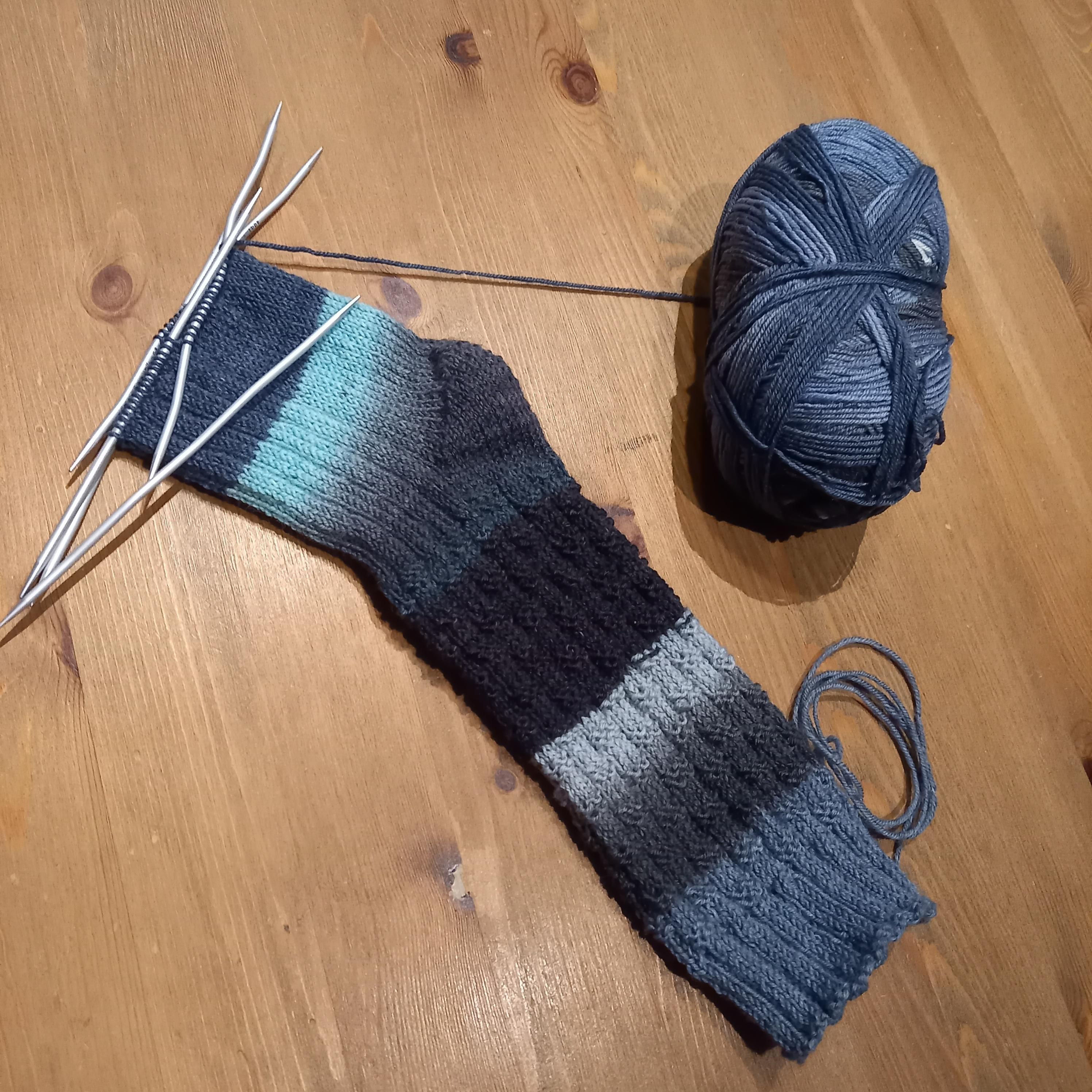 Eine selbstgestrickte, fast fertige Socke aus Wolle in verschiedenen Blautönen, es fehlt nur noch die Spitze.