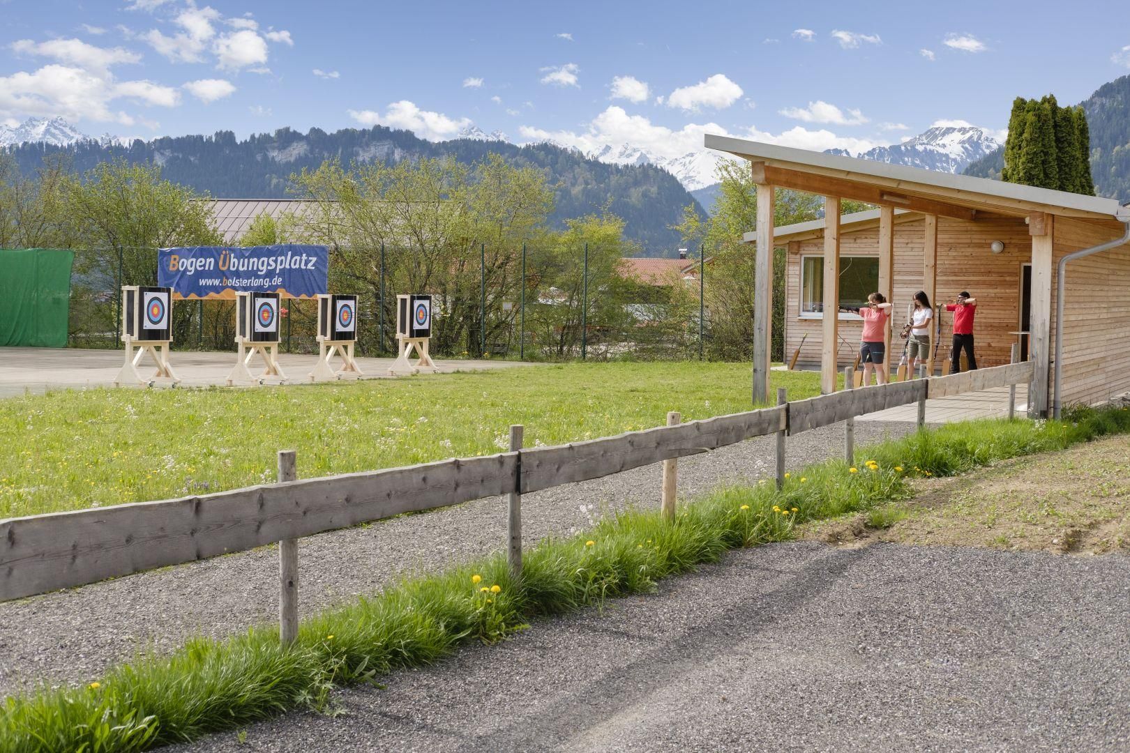Vier Zielscheiben auf grüner Wiese. Drei Personen mit Bögen unter hölzernem Unterstand.