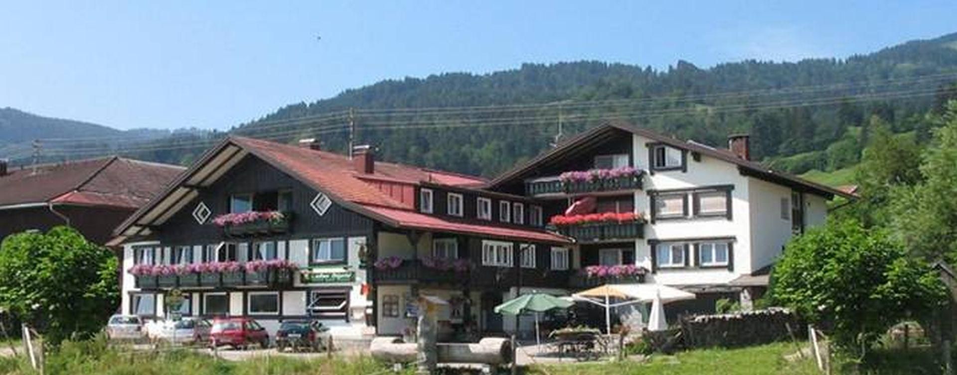 Dunkles Fachwerk, rote Fensterläden, pinke Blumen. Tische, Schirme außen. Autos, grüne Landschaft, Berge.