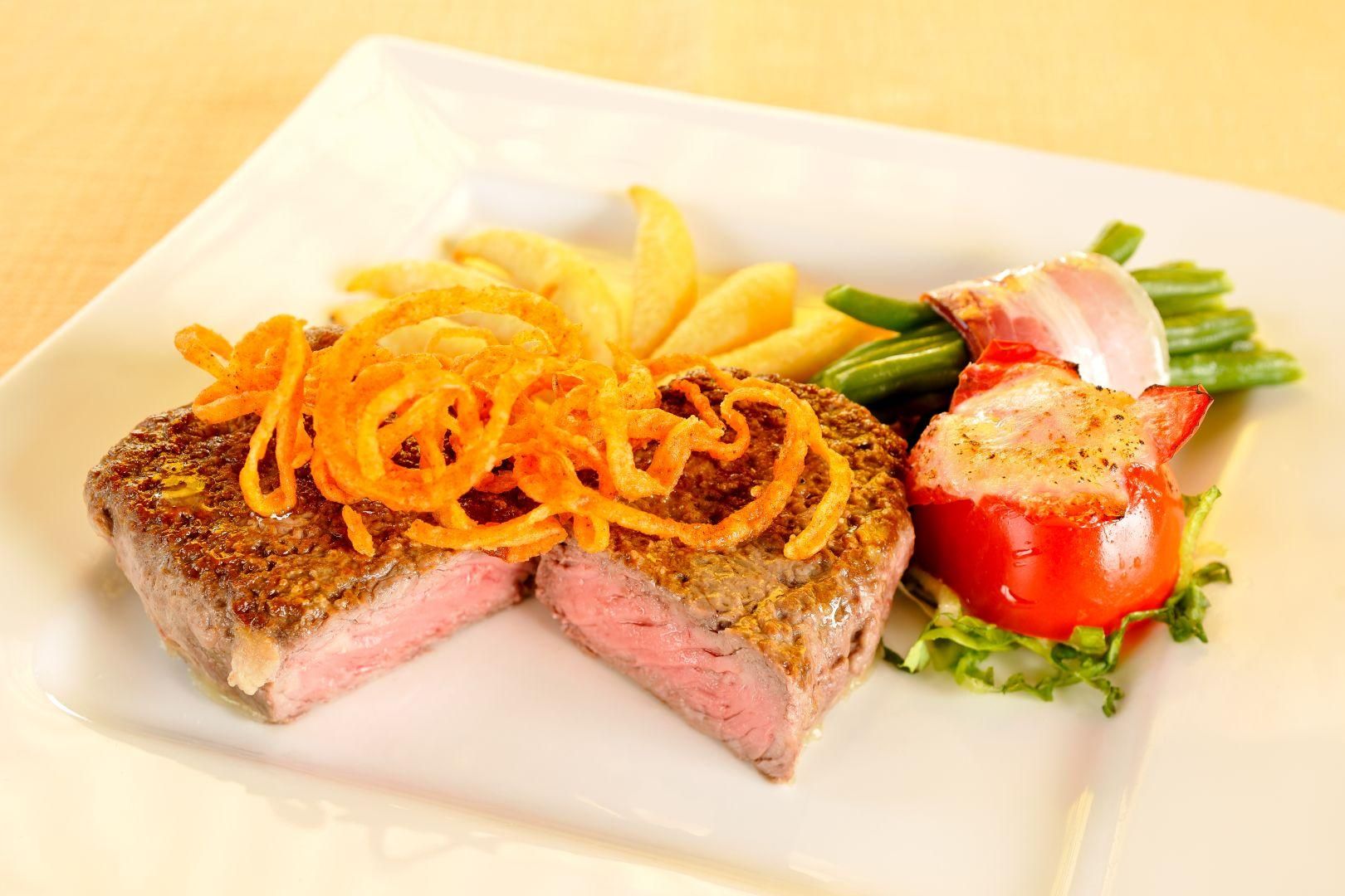 Ein zart rosa gebratenes Steak mit gerösteten Zwiebeln, einer Grilltomate und grünen Bohnen, mit Speck umwickelt.