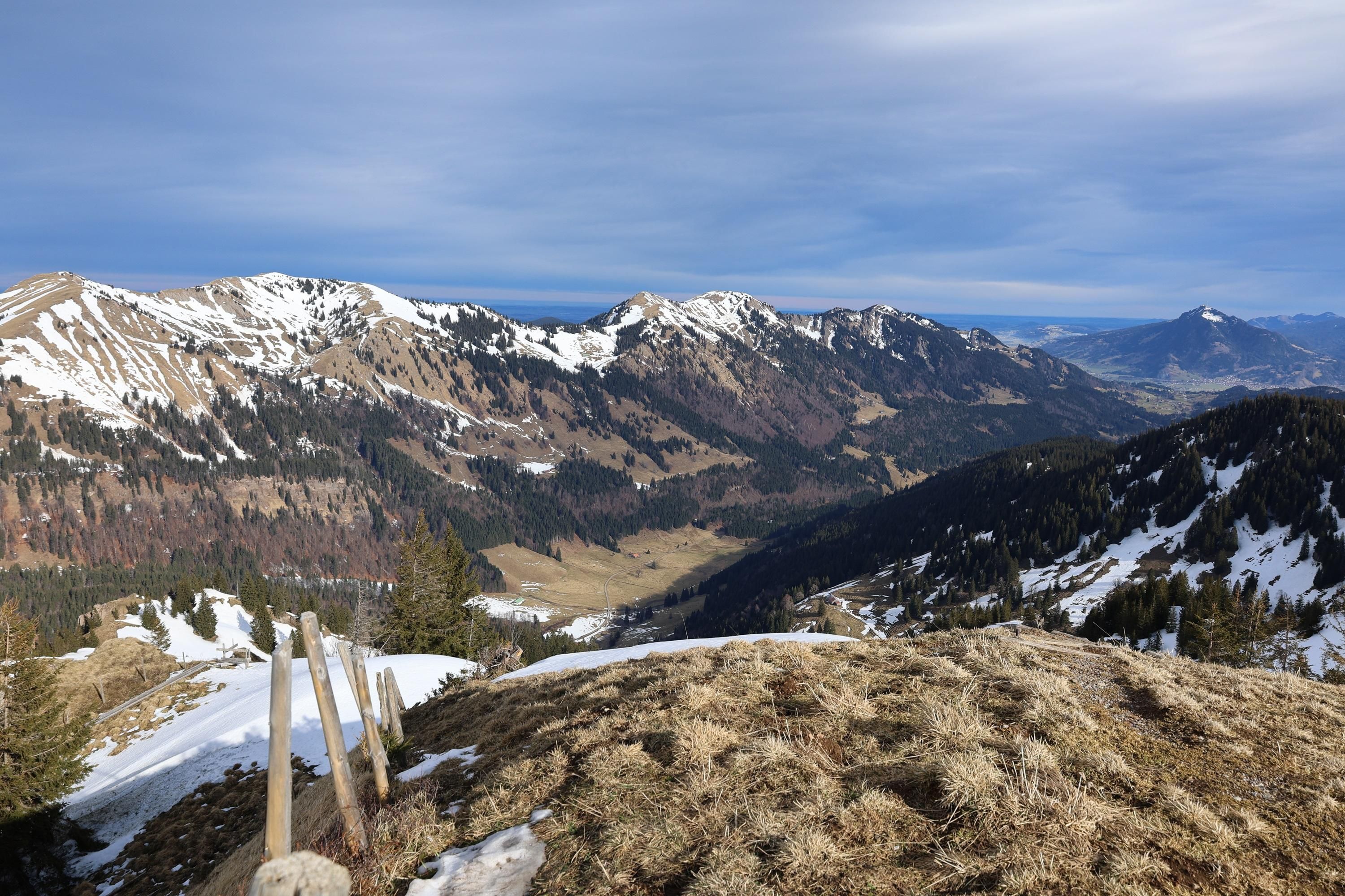 Eine malerische Winterlandschaft im Naturpark Nagelfluhkette mit schneebedeckten Bergen und Tälern unter einem bewölkten Himmel.