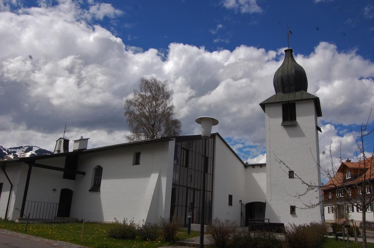Die Evangelische Kirche im Frühling, das Gras ist hellgrün, der Himmel blau mit großen Wolken.