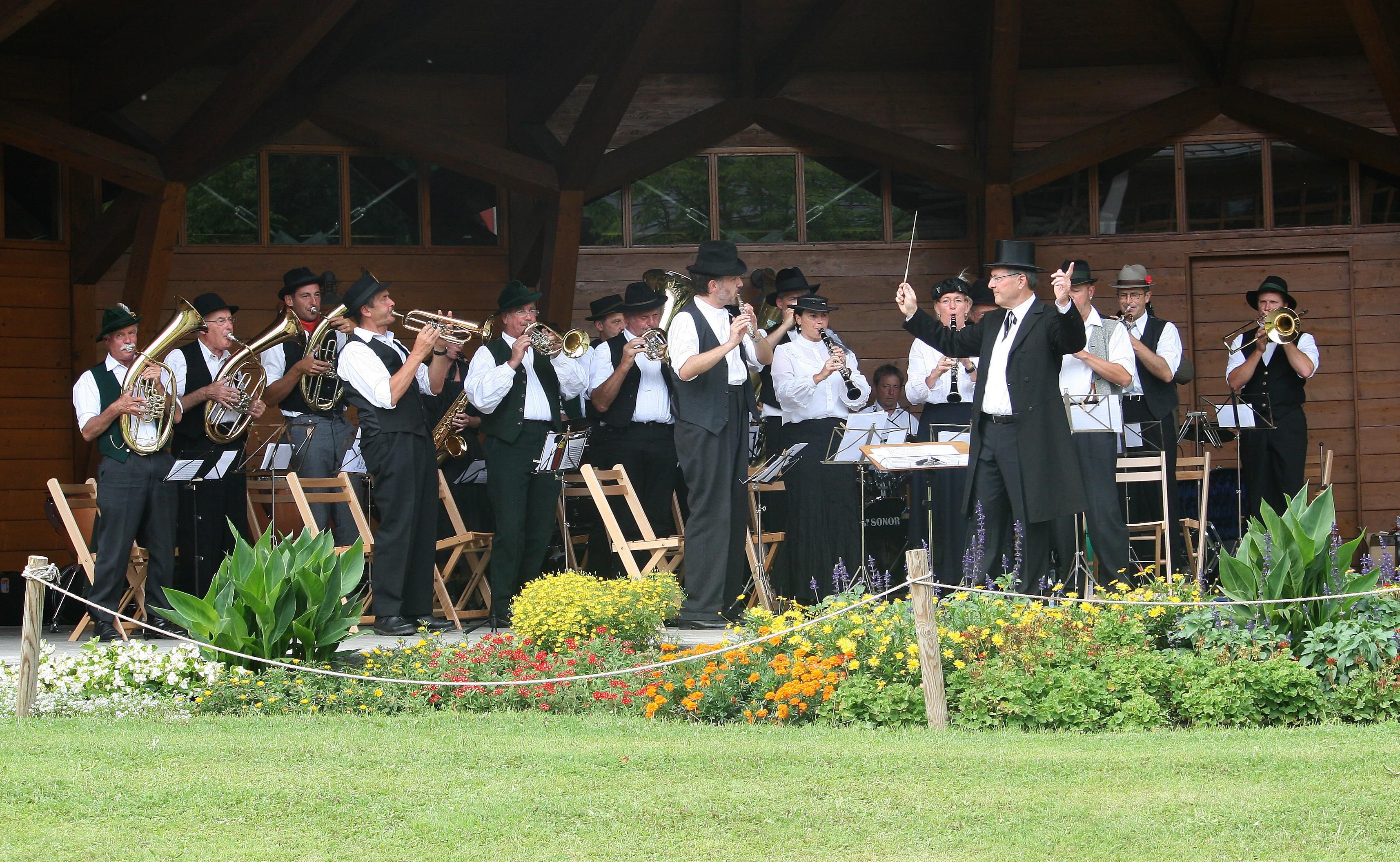 Blaskapelle "Üsrangierte" spielt auf einer Freilichtbühne. Dirigent in Zylinder und Anzug. Viele Blasinstrumente.