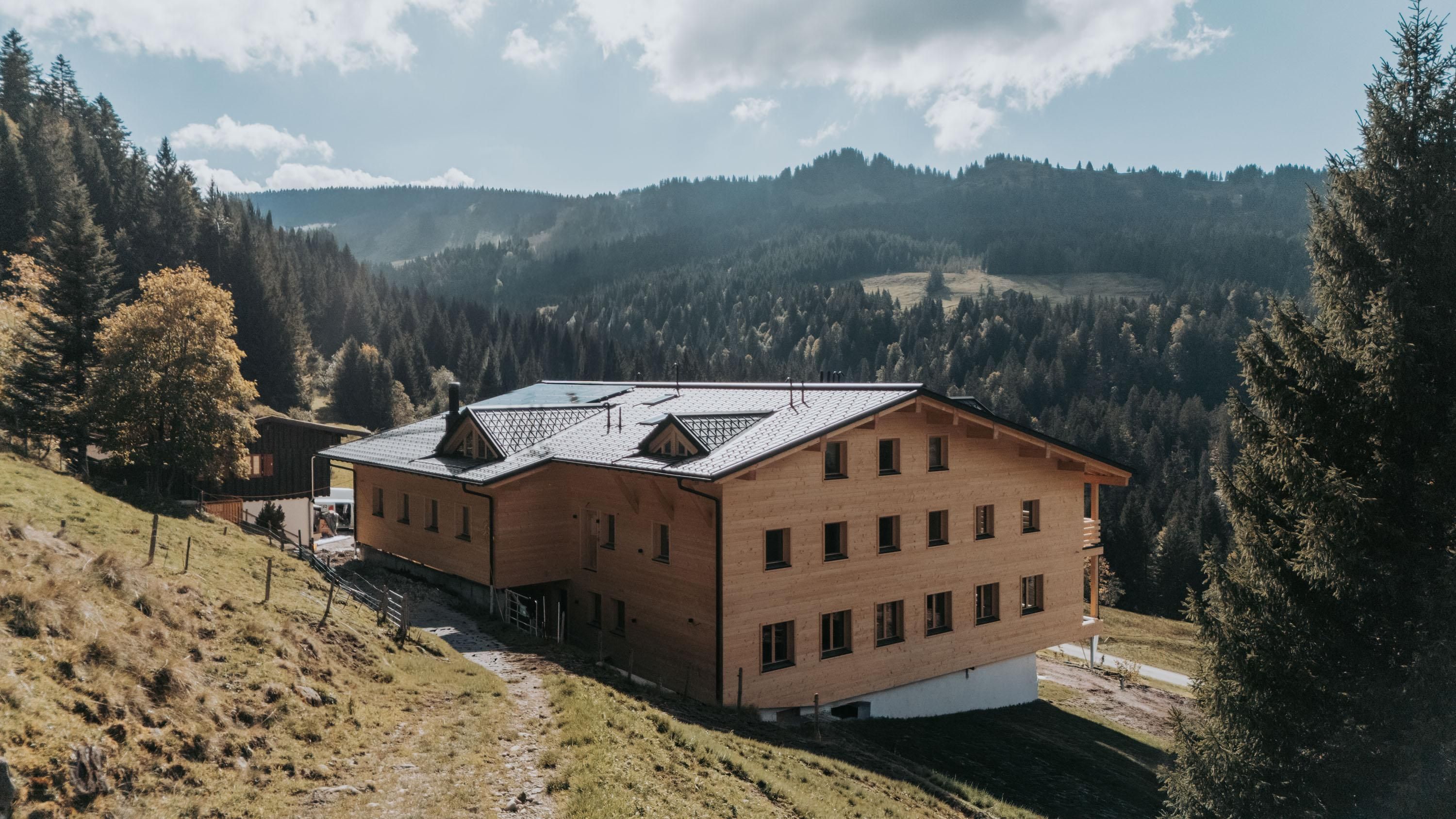 Ein großes, modernes Holzhaus steht allein auf einer sonnigen Bergwiese, umgeben von dichten Wäldern und sanften Hügeln. Im Hintergrund breitet sich eine weite Alpenlandschaft aus, die in weichem Licht unter einem leicht bewölkten Himmel liegt.