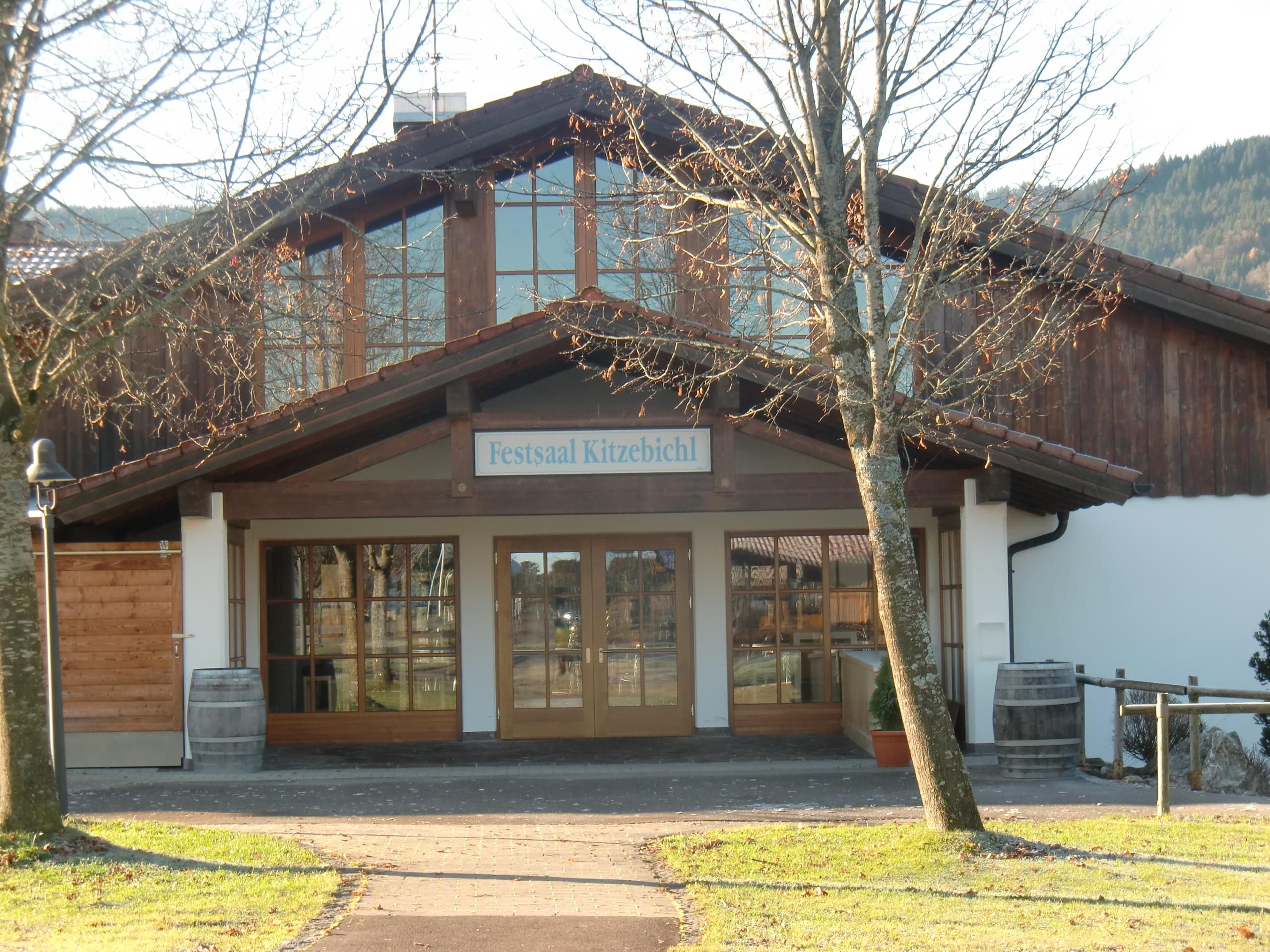 Eingang des Festsaals "Kitzebichl". Ein großes, holzverkleidetes Gebäude mit vielen Fenstern und einem braunen Ziegeldach.
