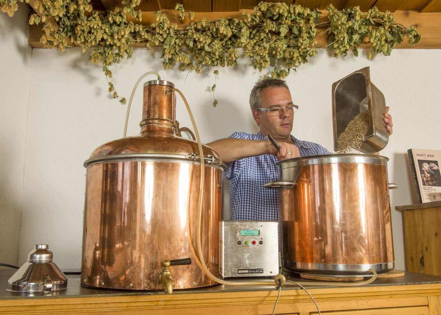 Mann braut Bier in Kupferkesseln, Hopfenranken hängen darüber, ein Buch liegt daneben.