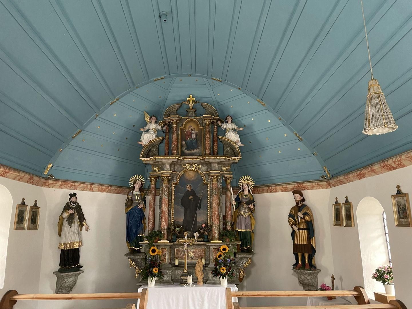 Innenansicht einer Kapelle mit blau gestrichener Decke und einem reich verzierten, goldenen Altar. Mehrere Heiligenfiguren stehen an den Seiten.