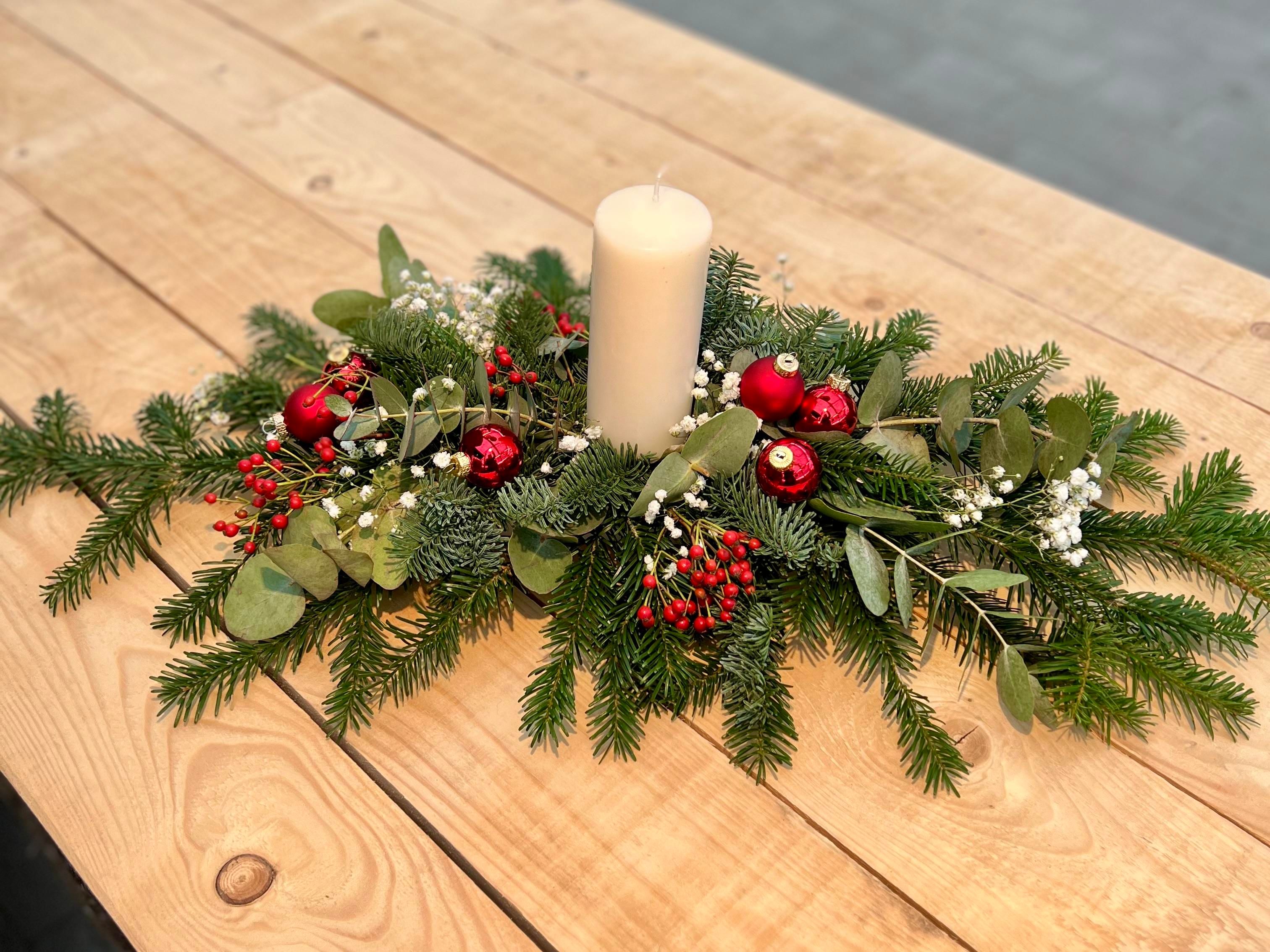 <p>Eine längliche, schmale Tischdekoration aus Tannen- und Eukalyptuszweigen mit roten Beeren und einer weißen Kerze.</p>