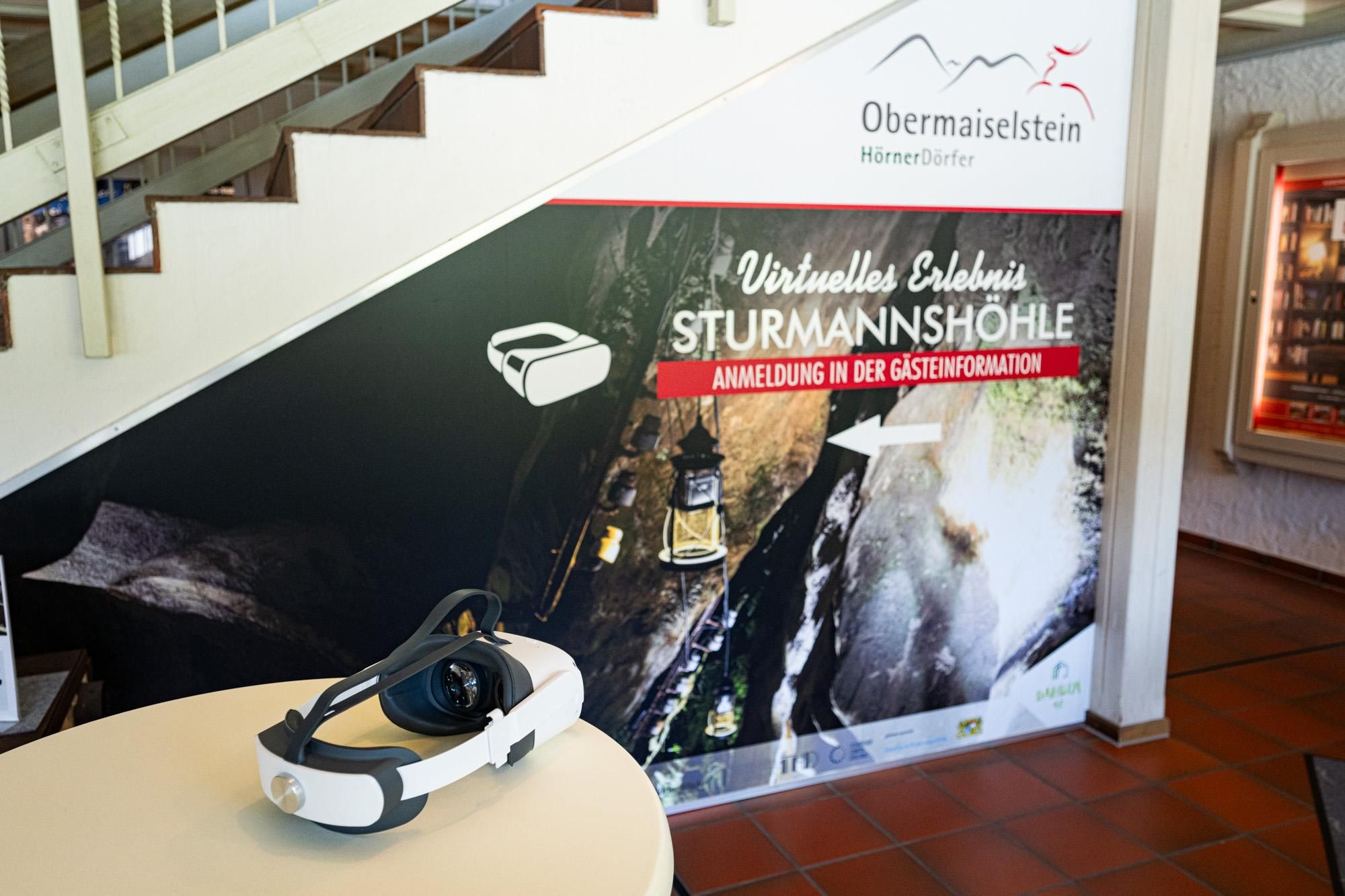 Tisch mit VR-Brille im Vordergrund. Wandplakat "Virtuelles Erlebnis STURMANNSHÖHLE, Anmeldung in der Gästeinformation".