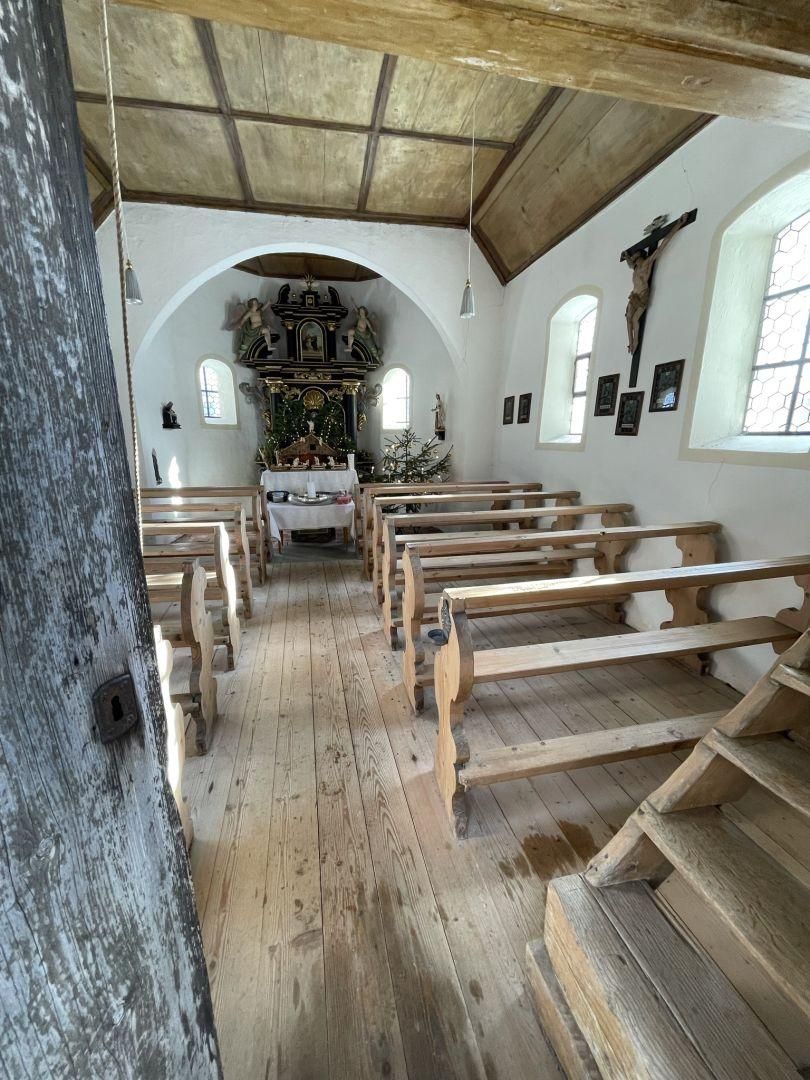 Innenansicht der Marienkapelle Kierwang. Helle Wände, Holzbänke und -decke. Kleiner, reich verzierter Altar. Kruzifix und Bilder an der Wand. Licht fällt durch die Fenster.