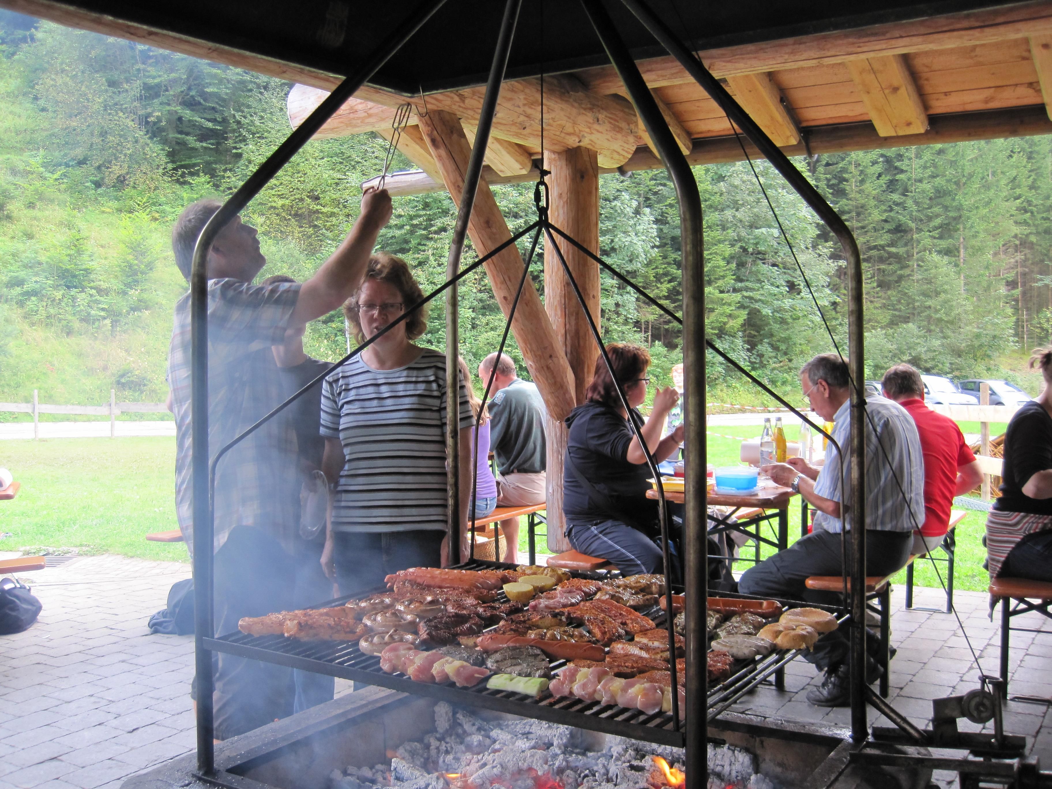 Auf einem großen Grill im Freien, unter einem Holzdach, brät eine Person Fleisch und Würste.