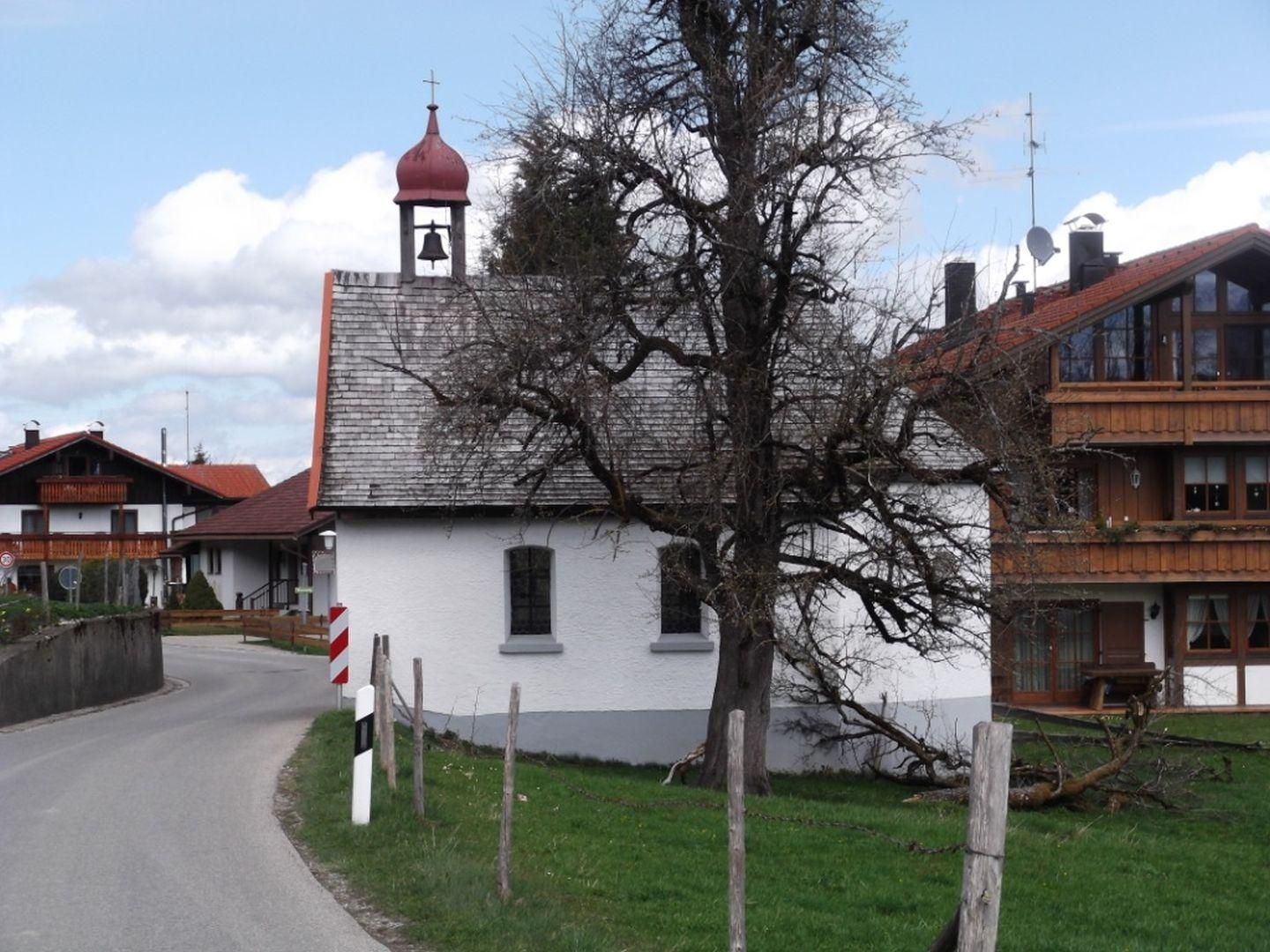 Weiße Marienkapelle Kierwang mit rotem Zwiebelturm und Glocke. Schiefergraues Dach. Kleine, dunkle Fenster. Steht an einer Straße mit Leitpfosten. Baum daneben.