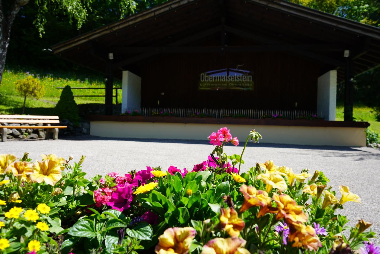 Ein großer Pavillon mit dunklem Holzdach und weißer Bühne, davor bunte Blumenbeete mit Petunien. Im Hintergrund Bäume und ein sonniger Himmel.