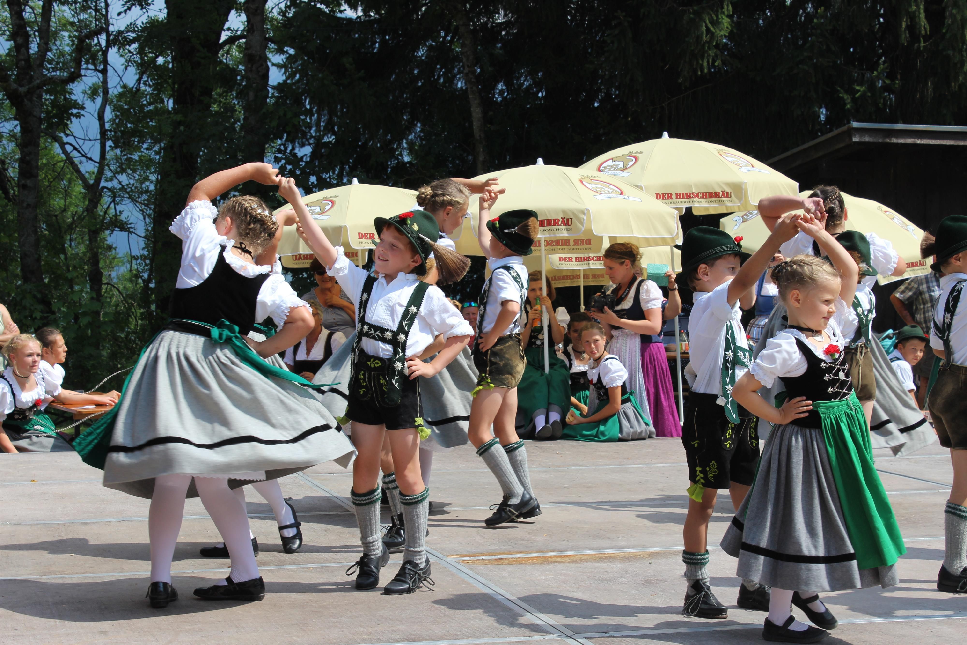 <p>Eine Gruppe junger Tänzerinnen und Tänzer in traditioneller bayerischer Tracht führt einen Volkstanz auf einer hellen Bühne im Freien auf.</p>
