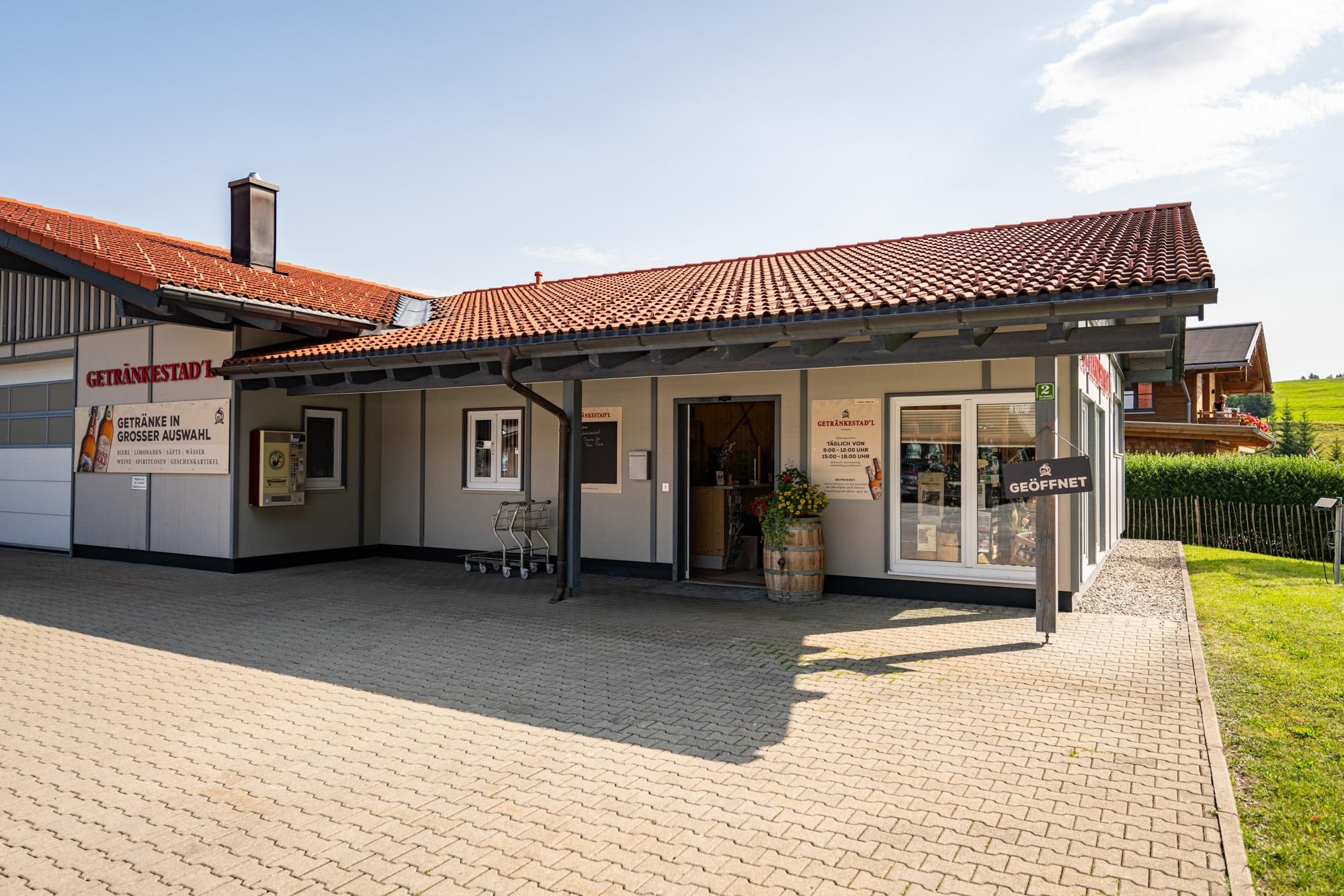Außenansicht des Getränkestadls Obermaiselstein. Helles, ebenerdiges Gebäude mit rotbraunem Ziegeldach und grauem Anbau.