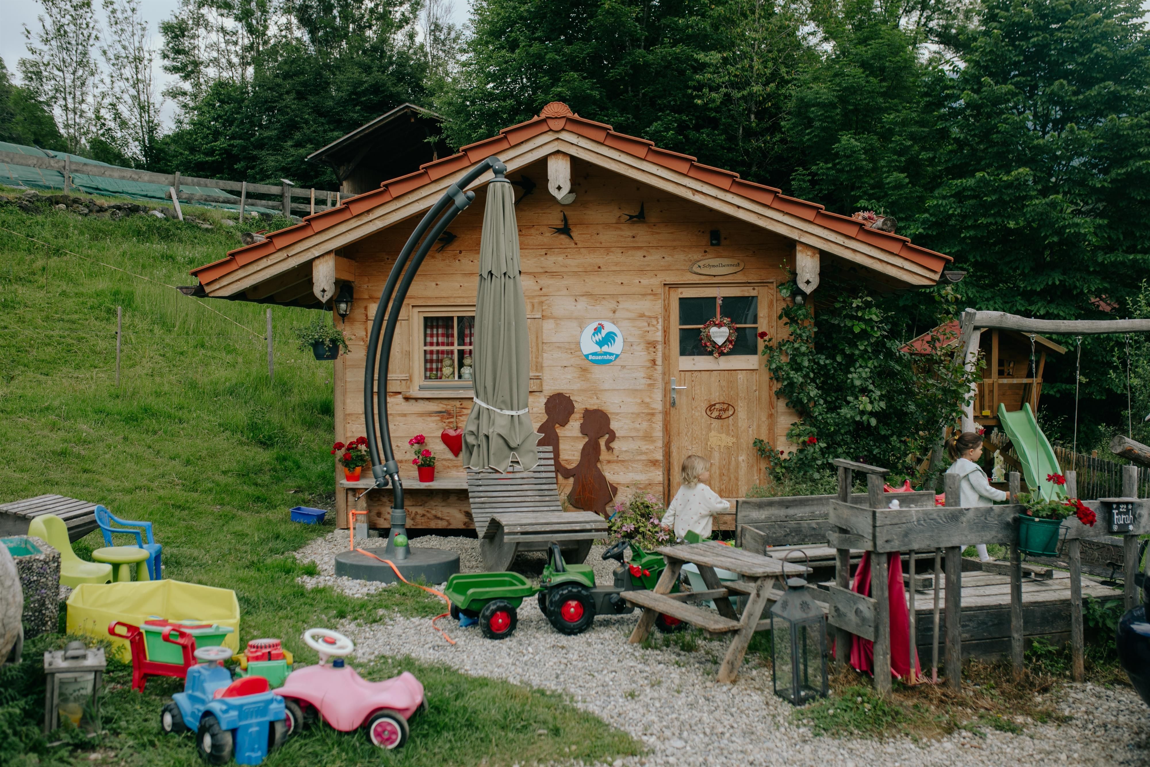 Spielhütte im Garten eines Bauernhofs in Fischen. Davor spielen Kinder mit Spielzeugautos, Rutsche und Sandkasten.