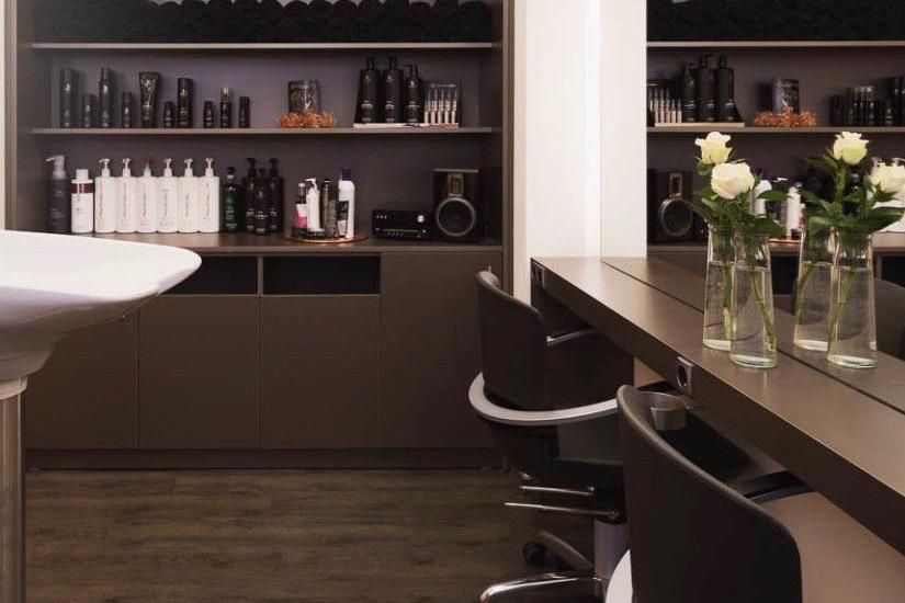 Friseursalon: Waschbecken links, Regale mit Produkten an der Wand. Rechts Spiegel mit Stühlen und Vasen mit weißen Rosen.