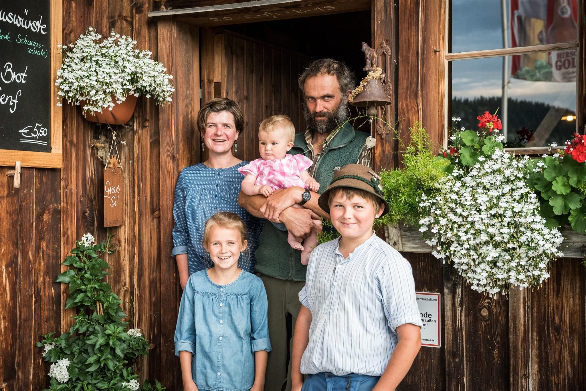 Familie Milz, die Gastgeber der Alpe Osterberg in Tiefenbach, stehen vor der offenen Tür ihrer hölzernen Hütte.