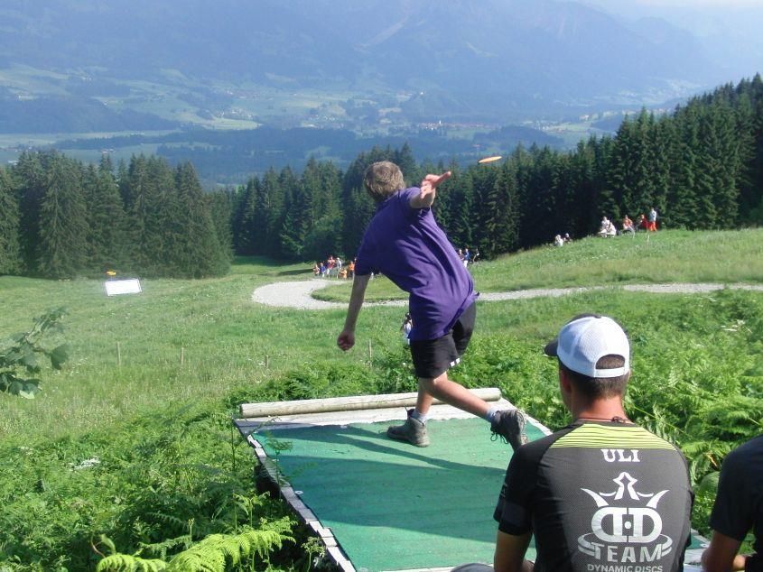 Ein Disc-Golfer wirft eine Scheibe von einer Matte über eine Bergwiese mit Wald und Tal.