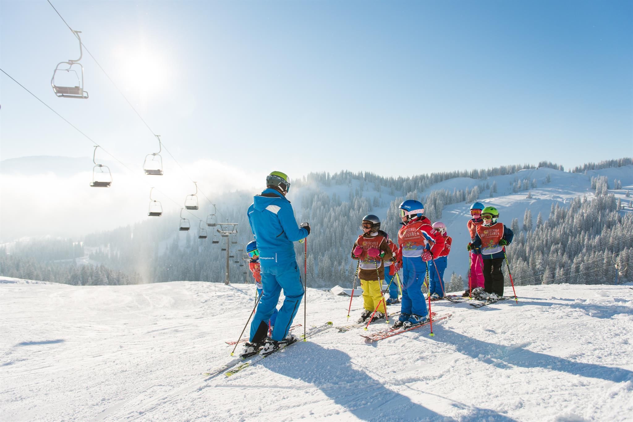 Ein schöner Wintertag mit blauem Himmel: ein Skilehrer erklärt den Kindern auf Ski etwas, alle tragen einen Helm.
