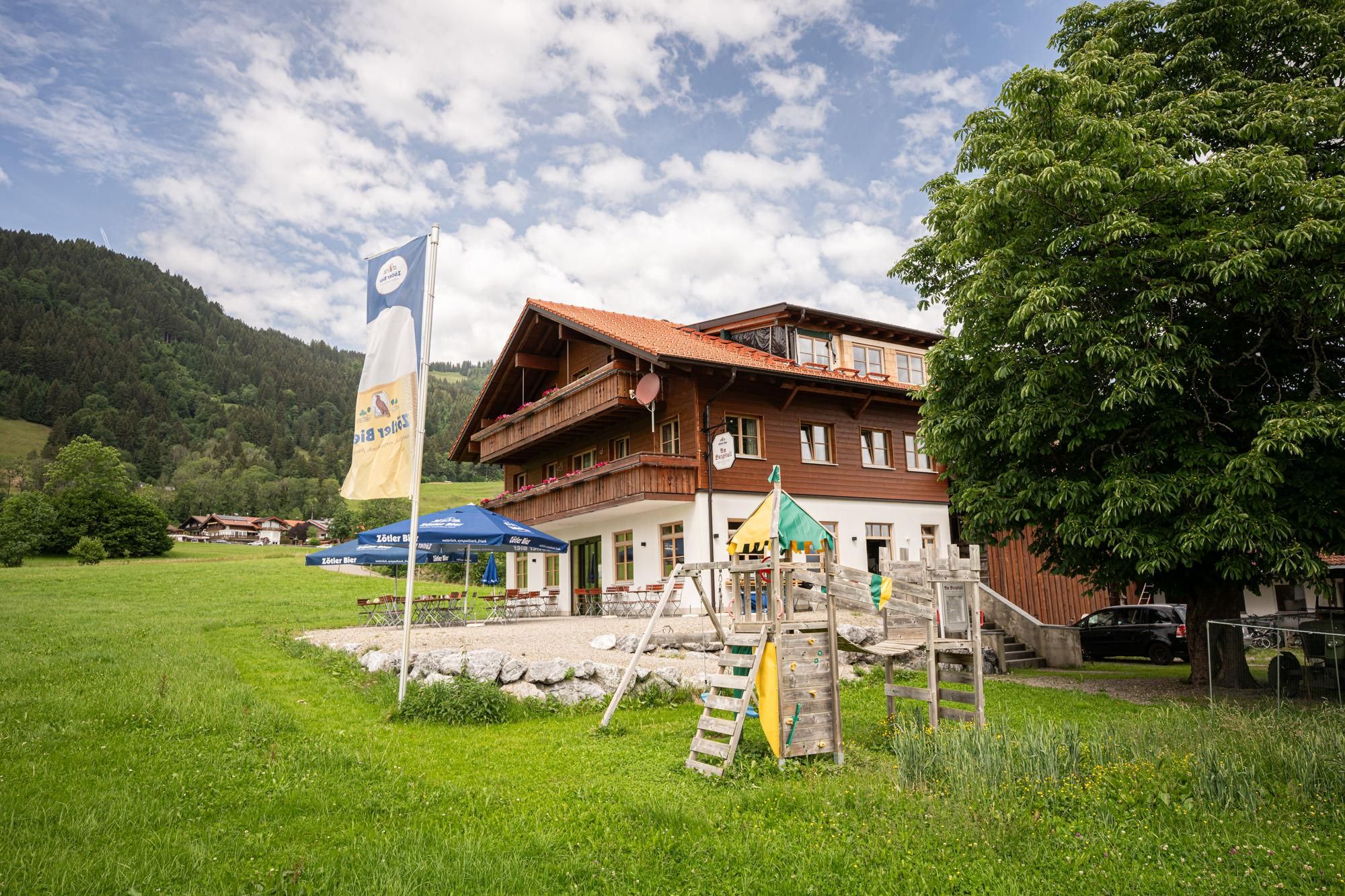 Ein traditionelles Allgäuer Bauernhaus umgeben von grünen Wiesen und Bäumen. Im Vordergrund sind Spielmöglichkeiten für Kinder zu sehen.